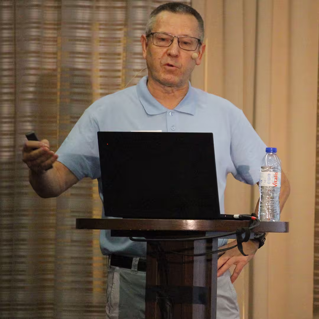 Morten Kromberg, CTO of Dyalog Ltd, stands at a wooden podium during Dyalog '22, delivering a presentation. He wears a light blue Dyalog polo shirt and light grey trousers, holding a presentation remote in one hand while gesturing with the other. A laptop sits open on the podium, with a bottle of water nearby. The background features beige curtains with soft vertical folds.