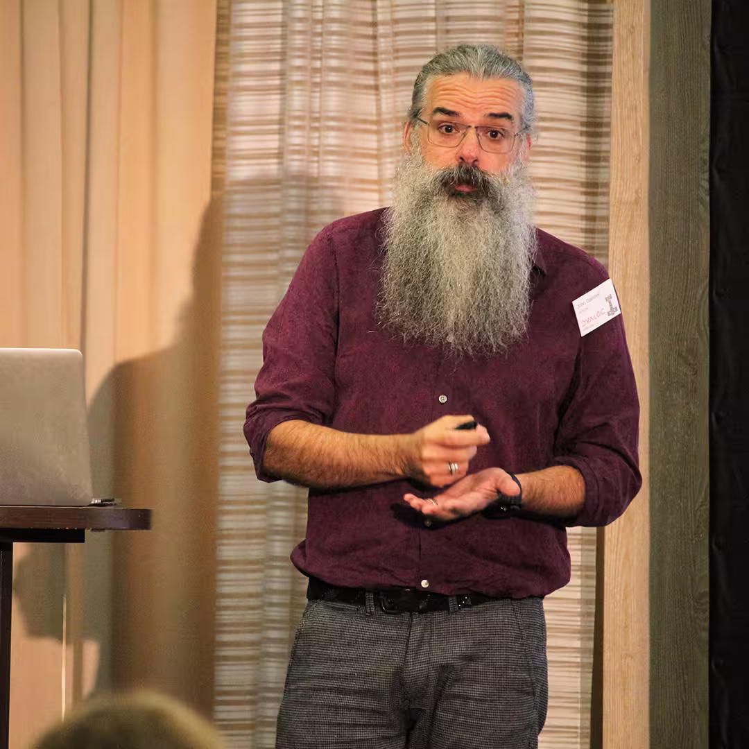 John Daintree, Chief Architect at Dyalog Ltd, stands on stage at Dyalog '22, delivering a presentation. He wears a burgundy shirt and grey trousers, holding a presentation remote in one hand while gesturing with the other. His long grey beard and glasses give him a distinctive appearance. A laptop sits on a nearby podium, and the background features beige curtains with soft vertical folds. The edge of an audience member's head is visible in the foreground.