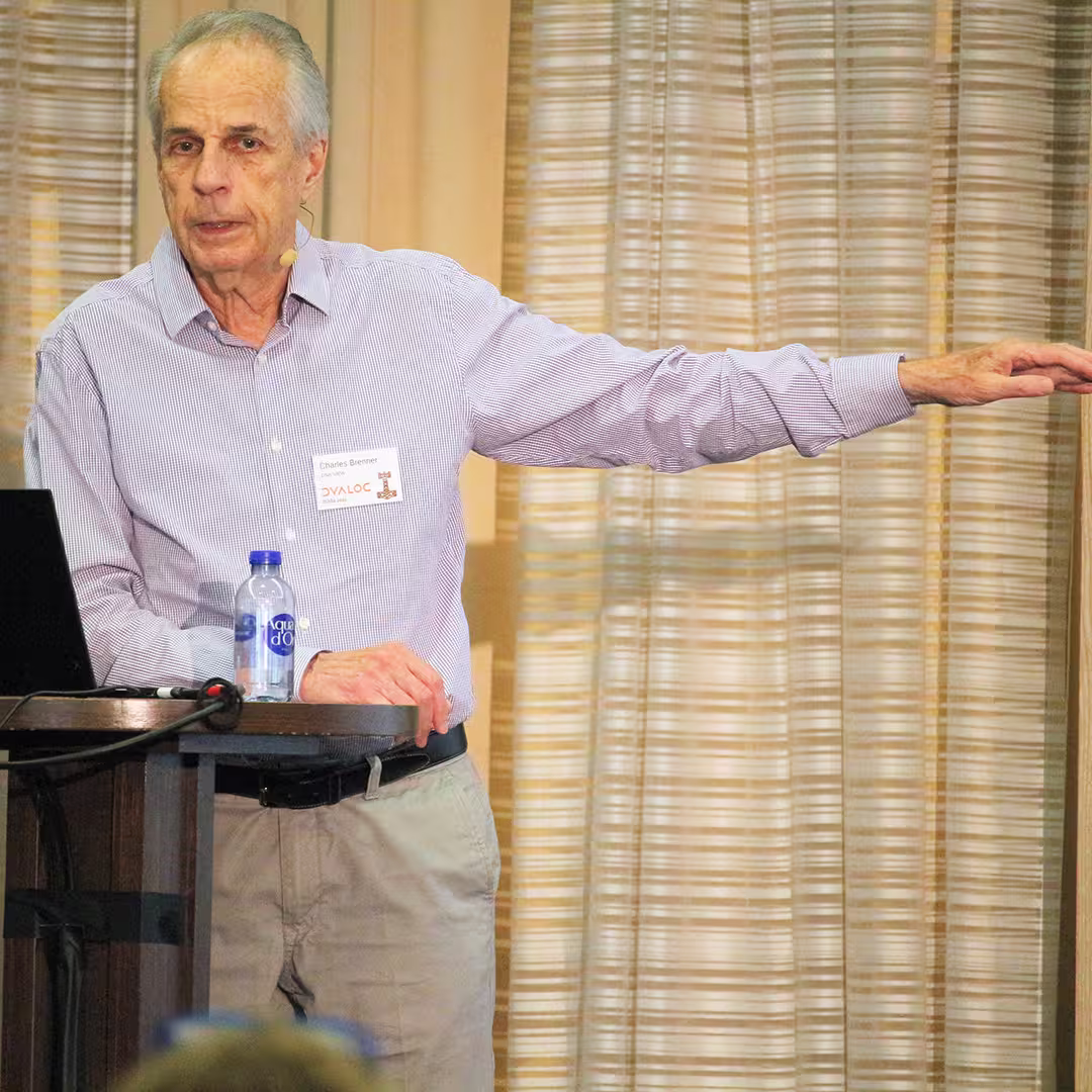 Charles Brenner stands at a wooden podium during Dyalog '22, delivering a presentation. He wears a light purple checkered shirt and light grey trousers, gesturing to the side with one arm extended. A laptop sits on the podium, alongside a bottle of water. The background features beige curtains with soft vertical folds. The edge of an audience member's head is visible in the foreground.