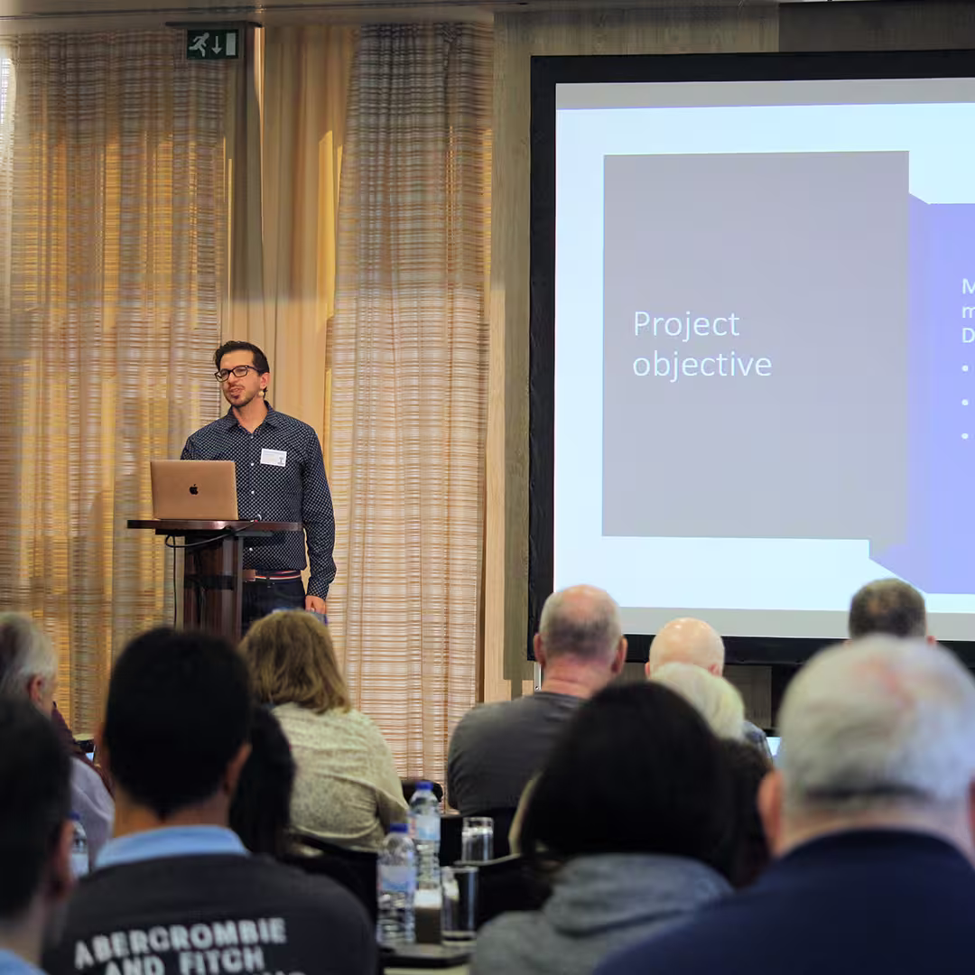 ilgamesh Athoraya of Tiamatica AB stands at a wooden podium during Dyalog '22, delivering a presentation. He wears a dark blue, patterned shirt and glasses, speaking into a headset microphone. A laptop sits open on the podium. Behind him, a large screen displays a slide titled "Project objective." The background features beige curtains with soft vertical folds, and the heads of several audience members are visible in the foreground.