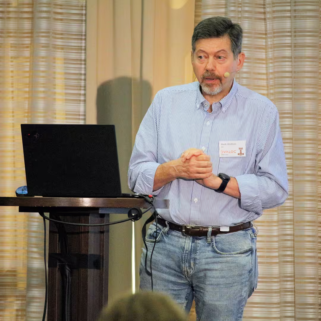 Mark Wolfson, CTO at BIG, consultants to the jewellery industry, stands at a wooden podium during Dyalog '22, delivering a presentation. He wears a light blue striped shirt and jeans, speaking into a headset microphone while clasping his hands together. A laptop sits open on the podium. The background features beige curtains with soft vertical folds, and the edge of an audience member's head is visible in the foreground.