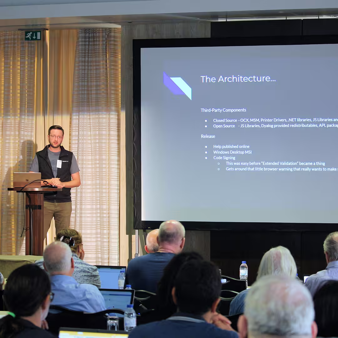 Norbert Jurkiewicz of The Carlisle Group stands at a wooden podium during Dyalog '22, delivering a presentation. He wears a black vest over a grey shirt and glasses, speaking into a headset microphone while standing beside a laptop. Behind him, a large screen displays a slide titled "The Architecture," listing third-party components and release details. The background features beige curtains with soft vertical folds, while several audience members, some with laptops open, are visible in the foreground.