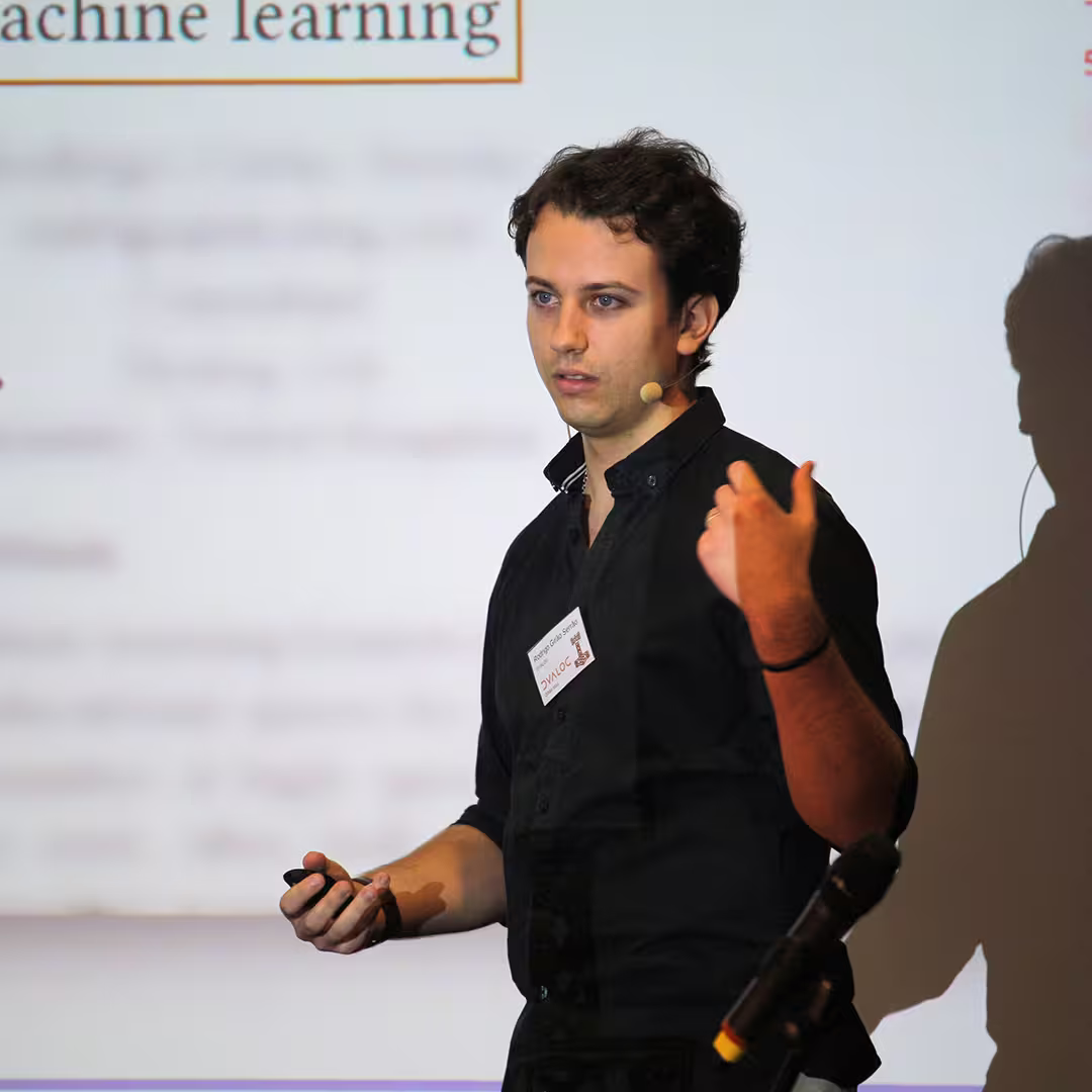 Rodrigo Girão Serrão of Dyalog Ltd delivers a presentation at Dyalog '22. He wears a black shirt and speaks into a headset microphone while gesturing with one hand and holding a presentation remote in the other. Behind him, a slide with the words "machine learning" is projected onto the screen, casting his shadow onto the background. A microphone on a stand is visible in the foreground.