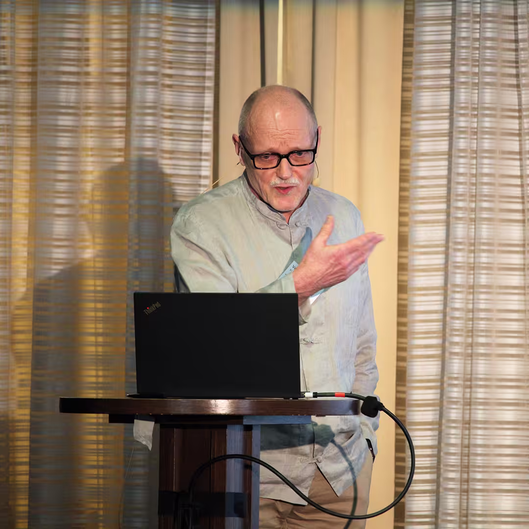 Veli-Matti Jantunen of Statistics Finland stands at a wooden podium during Dyalog '22, delivering a presentation. He wears a light grey, buttoned-up shirt and glasses, speaking into a headset microphone while gesturing with one hand. A laptop sits open on the podium, with connecting cables visible. The background features beige curtains with soft vertical folds.