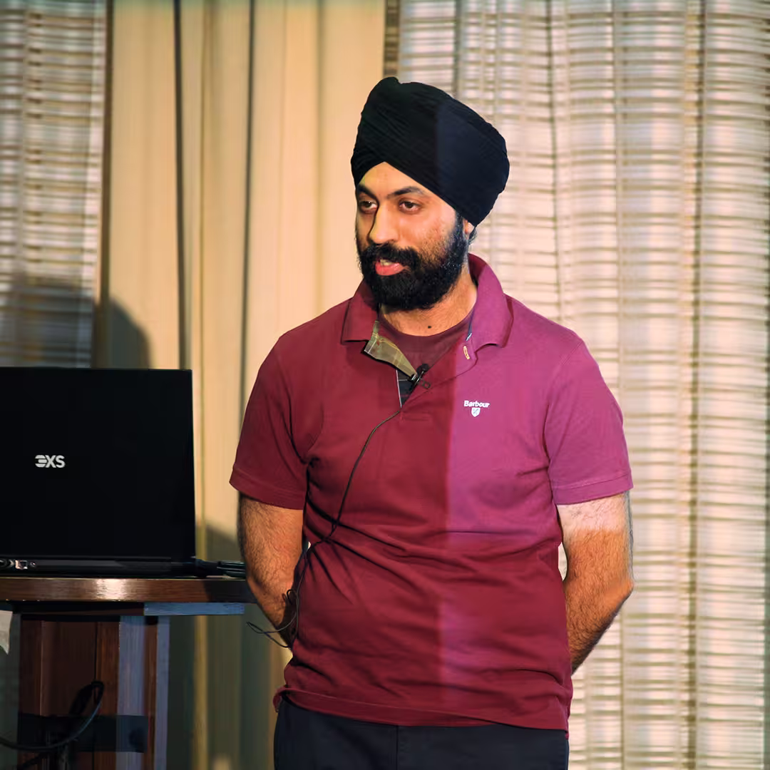 Karta Kooner of Dyalog Ltd stands on stage at Dyalog '22, delivering a presentation. He wears a burgundy Barbour polo shirt and a black turban, speaking into a headset microphone with his hands clasped behind his back. A laptop sits on a nearby wooden podium. The background features beige curtains with soft vertical folds.