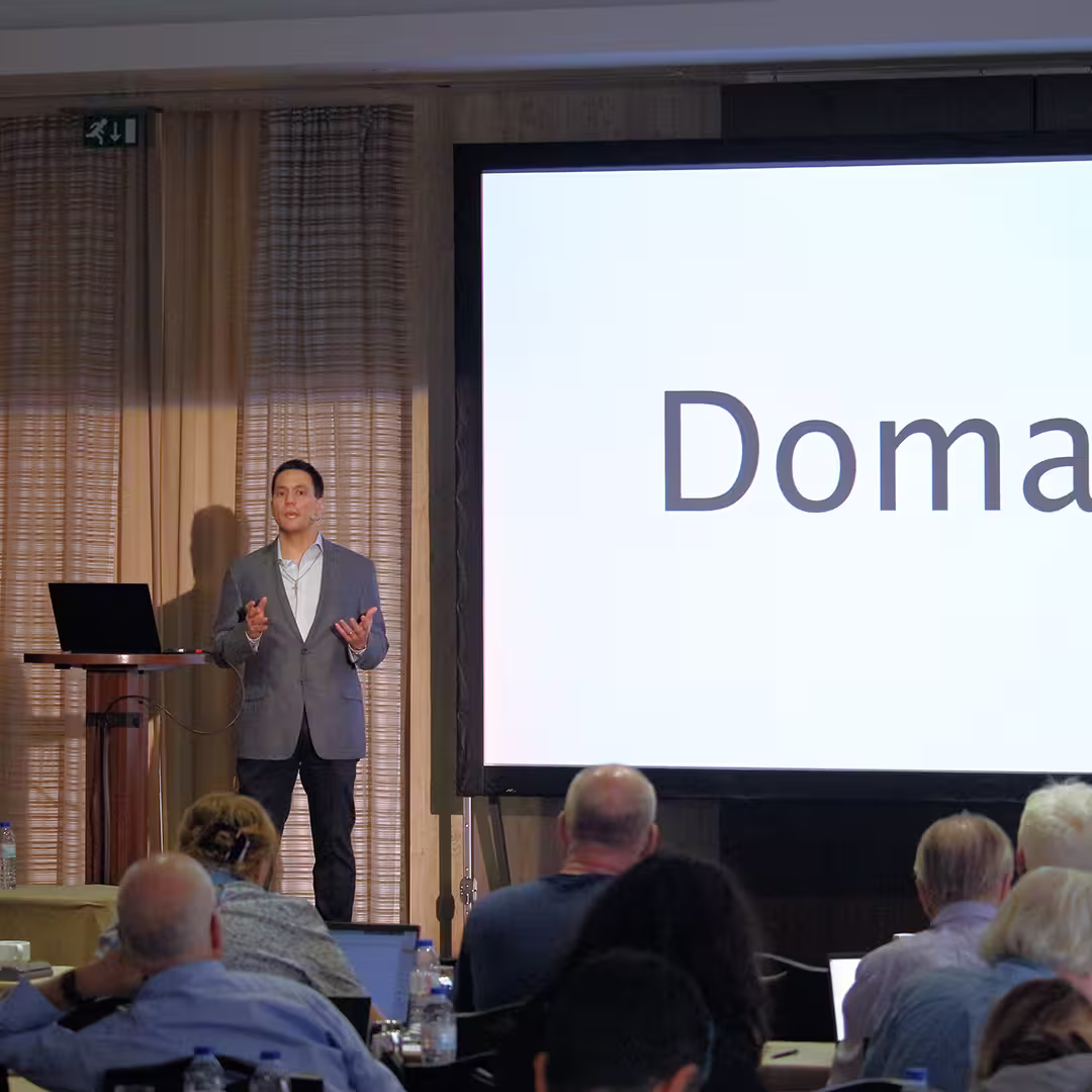Aaron Hsu stands on stage at Dyalog '22, delivering a presentation. He wears a grey blazer, white shirt, and dark trousers, gesturing with both hands while speaking into a headset microphone. A large screen behind him displays the word "Domain" in bold black letters on a white background. A laptop sits on a wooden podium to his left. Several audience members, some using laptops, are visible in the foreground. The background features beige curtains with soft vertical folds.