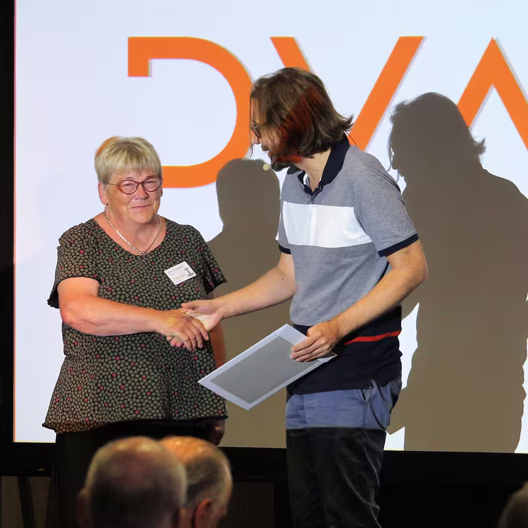 Gitte Christensen shakes hands with Michael Higginson on stage at Dyalog '22. Michael is receiving an award for winning the APL Problem Solving Competition (Professional Entrant). Gitte wears a dark green blouse with a pink polka-dot pattern, while Michael wears a grey and navy colour-blocked polo shirt. Behind them, a large screen displays the letters "DYA" in bold orange. Audience members are visible in the foreground.