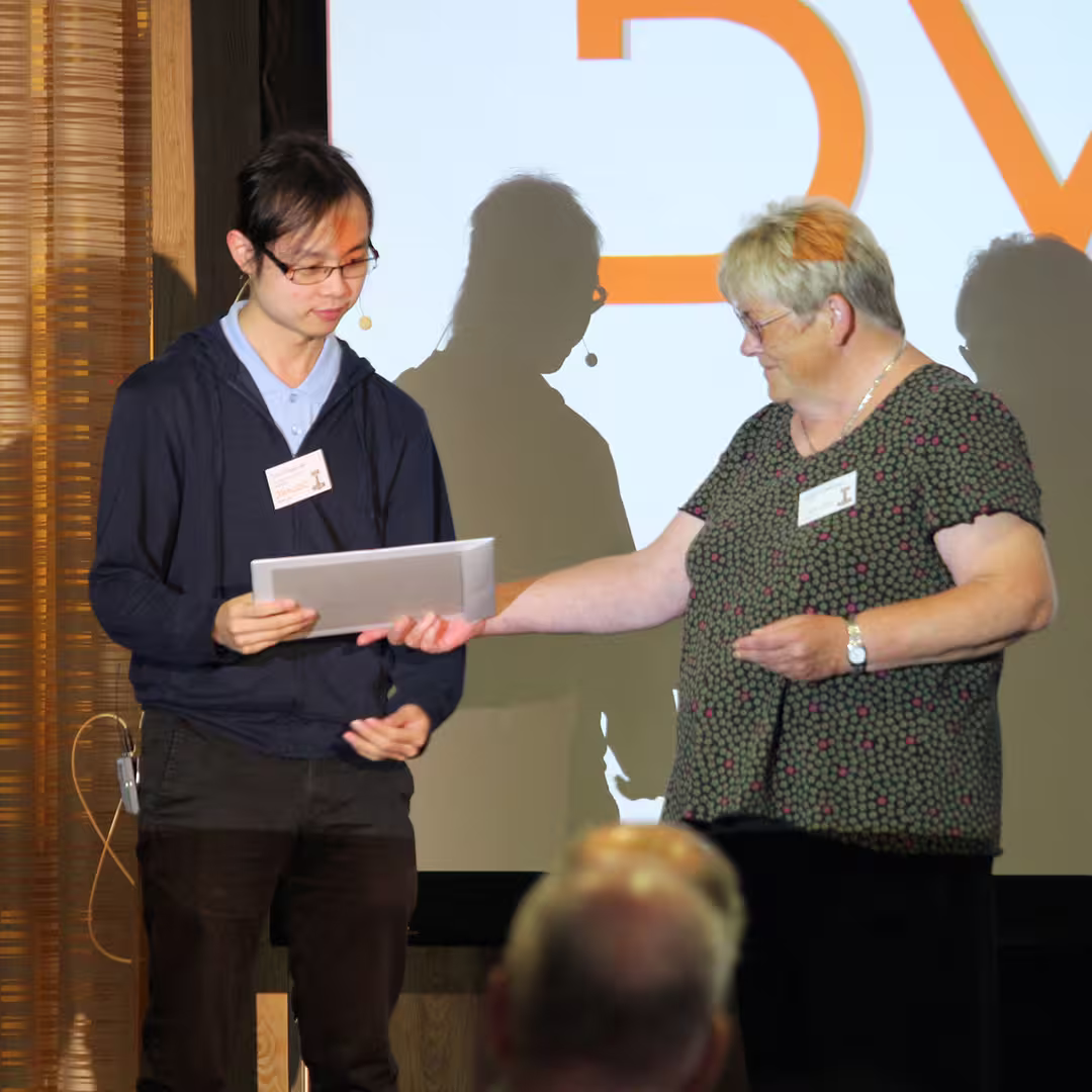 Gitte Christensen hands an award to Tzu-Ching Lee on stage at Dyalog '22 for winning the APL Problem Solving Competition. Gitte wears a dark green blouse with a pink polka-dot pattern, while Tzu-Ching wears a light blue shirt under a navy hoodie. Behind them, a large screen displays the letters "DYA" in bold orange. Audience members are visible in the foreground.