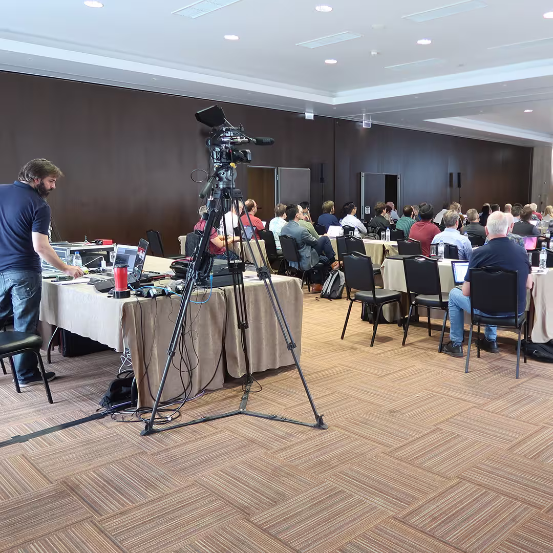 A conference session at Dyalog '22 shows delegates seated at tables, facing the front of the room. Each table is covered with beige tablecloths and set with water bottles, notepads, and laptops. In the foreground, a professional video camera stands on a tripod, aimed at the stage. To the left, Jason Rivers manages the AV setup from a table cluttered with cables, laptops, and equipment. The room features dark wood-panelled walls, a patterned carpet, and a white ceiling with recessed lighting.