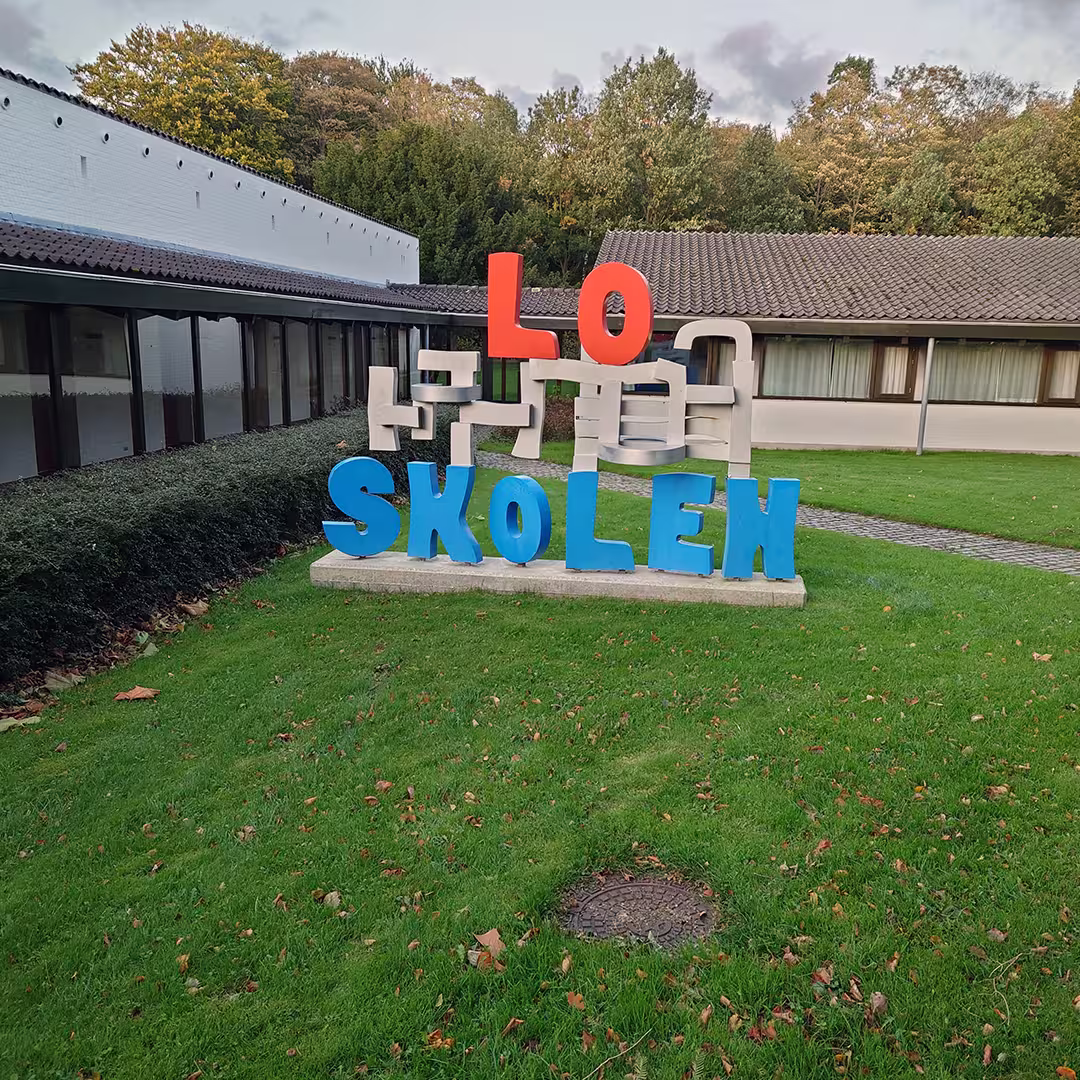 A colorful outdoor sign at the Konventum Convention Centre in Elsinore, Denmark, featuring the words "LO SKOLEN" in large, bold letters. "LO" is in red, while "SKOLEN" is in bright blue. Between the words, abstract beige shapes resembling interconnected blocks form part of the design. The sign is set on a concrete base on a well-maintained lawn with a few fallen leaves. Behind it, a low, white building with dark-tiled roofs and large windows is visible, with green trees in the background.