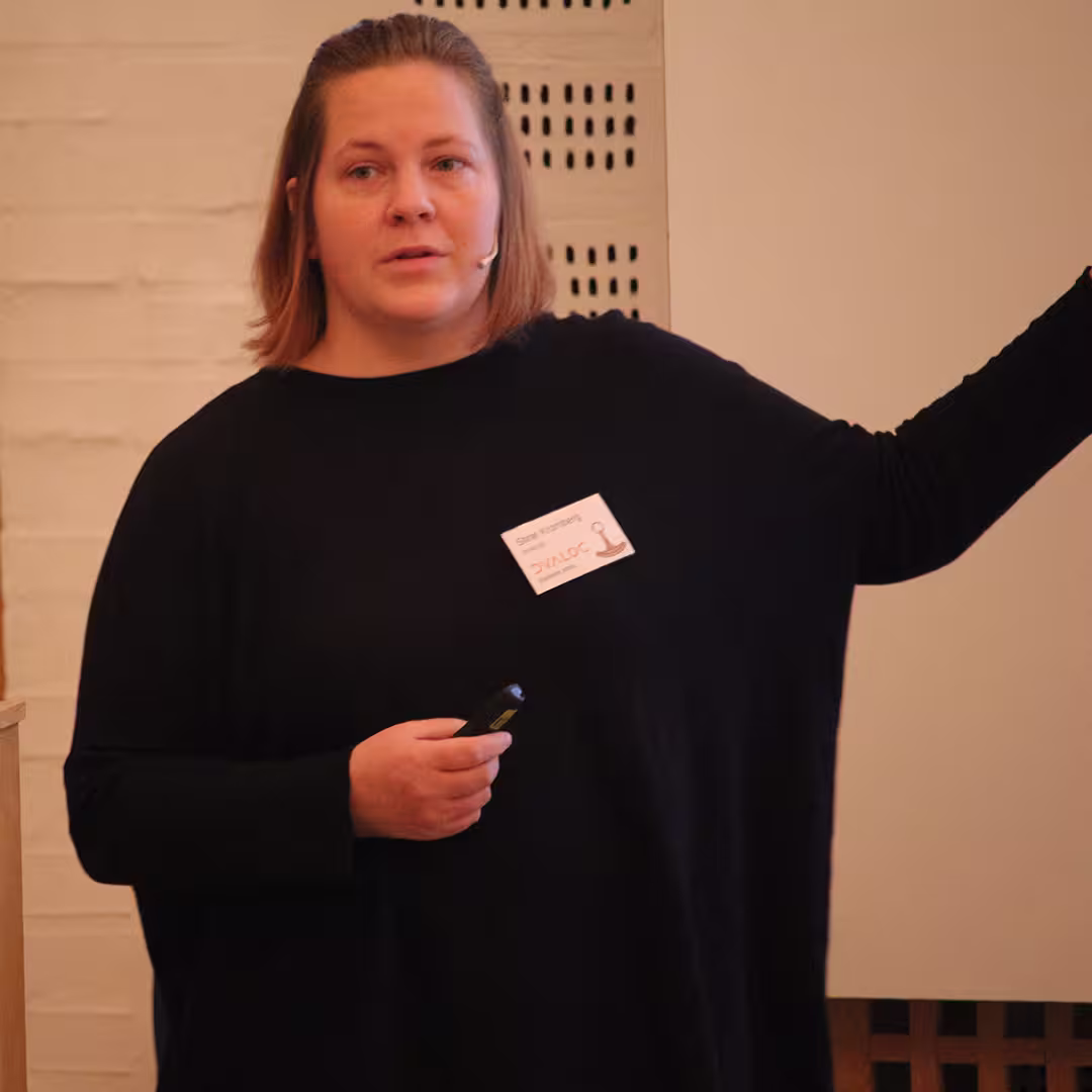 Stine Kromberg, wearing a black sweater and a name badge, delivers a presentation at Dyalog '23. She speaks into a headset microphone while gesturing towards a screen with her right hand. In her left hand, she holds a presentation remote. The background features a white brick wall and a section of the projector screen.