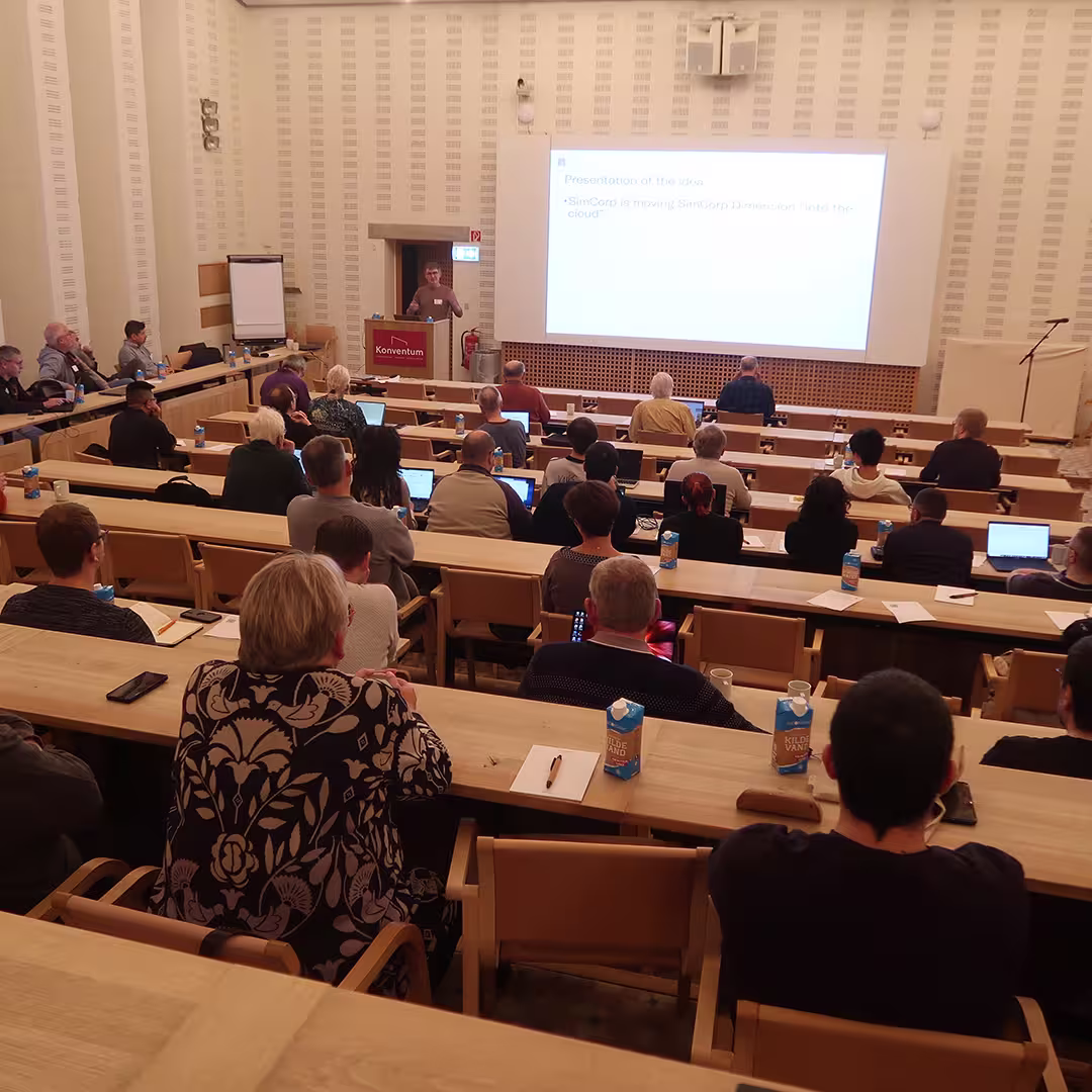 A speaker at Dyalog '23 stands at the podium in a tiered lecture hall, delivering a presentation. The large screen behind them displays a slide titled "Presentation of the idea," mentioning SimCorp moving to the cloud. Attendees sit at wooden desks, some taking notes or working on laptops, while others listen attentively. Many have cartons of water on their desks. The hall features warm lighting, wooden paneling, and a minimalist design, creating a focused yet comfortable setting.