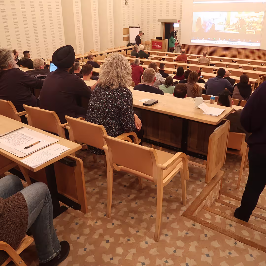 Attendees at Dyalog '23 sit in a tiered lecture hall, watching a presentation that includes a live video call displayed on a large screen at the front. The audience, seated at wooden desks with notepads and cups of coffee, listens attentively. Some take notes, while others lean forward in their chairs. A speaker stands at a podium near the screen, with another person assisting. The room has warm lighting, wooden accents, and patterned carpeting, contributing to a comfortable and professional atmosphere.