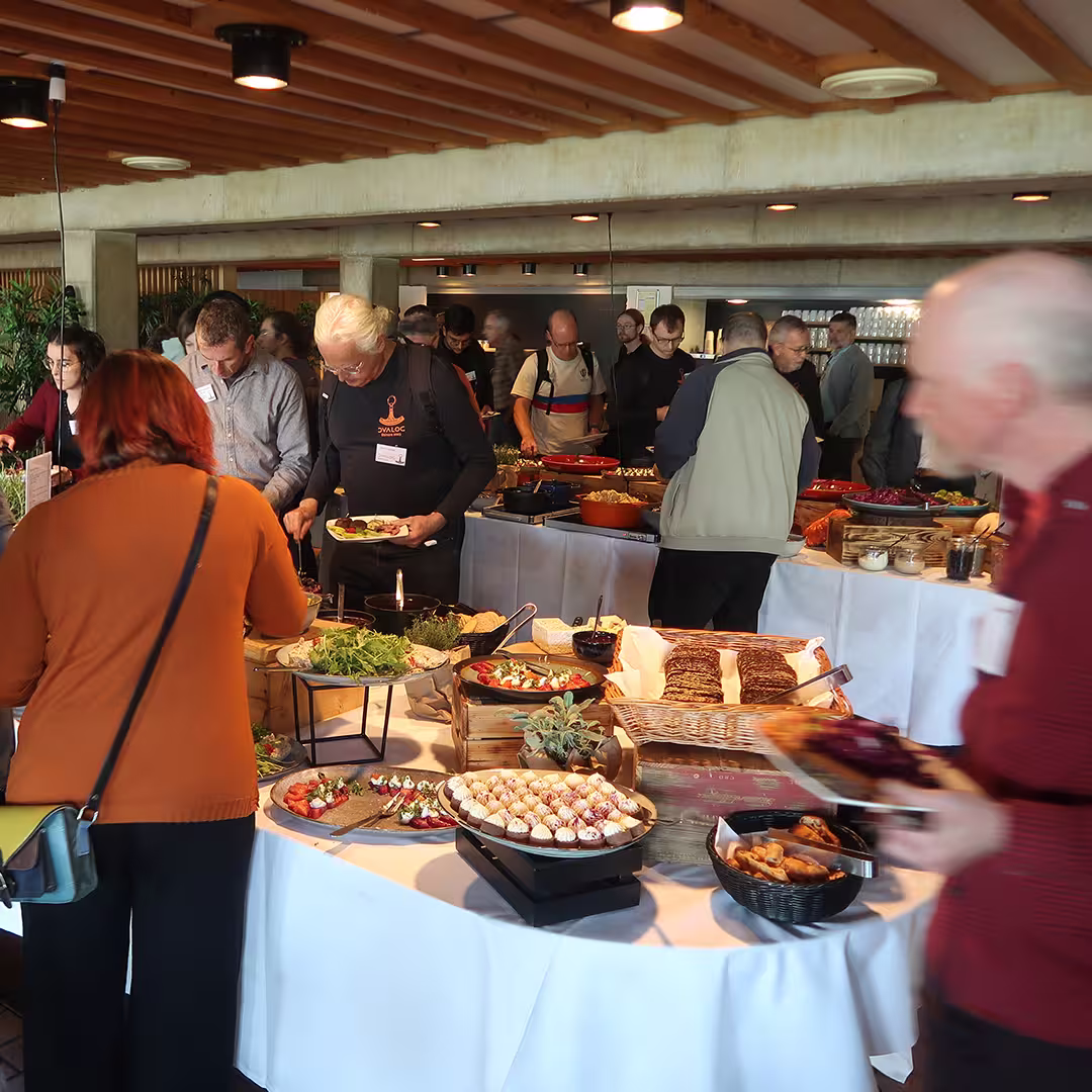 Attendees at Dyalog '23 gather around a buffet table filled with a variety of dishes, including salads, bread, pastries, and desserts. A man in the foreground, wearing a black Dyalog-branded long-sleeve shirt, is selecting food for his plate. Others are engaged in conversation while serving themselves. The dining area features wooden beams on the ceiling, warm lighting, and a modern, open kitchen in the background with stacked glassware and serving dishes.