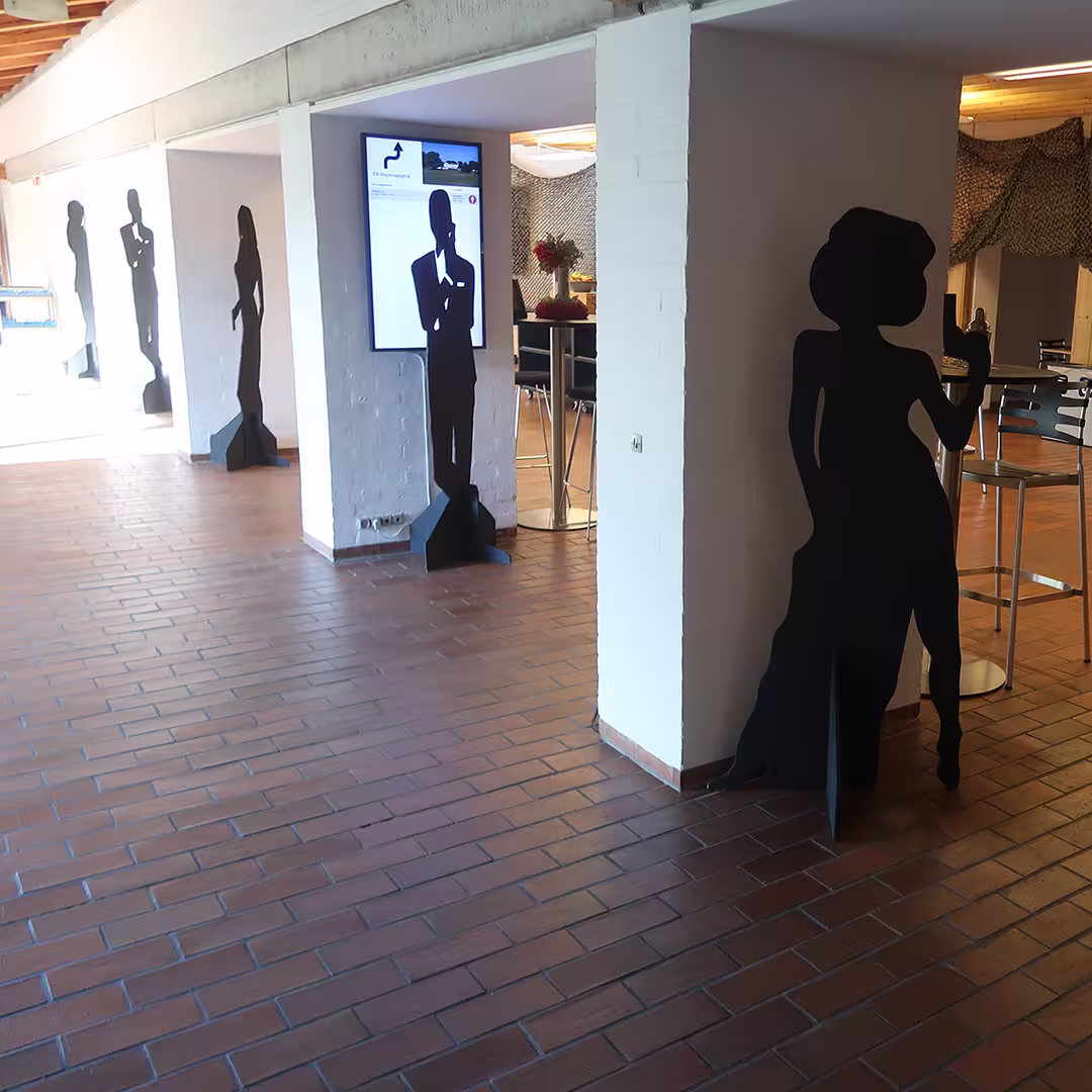A hallway at the Konventum Convention Centre in Elsinore, Denmark, decorated with black silhouette cutouts of elegantly dressed figures, evoking a James Bond or espionage theme. One silhouette in the foreground depicts a woman holding a cocktail glass, while others are positioned along the corridor. A digital screen displaying an event-related image is mounted on a white pillar. In the background, a lounge area with high tables and chairs is visible, with a vase of flowers adding a decorative touch.