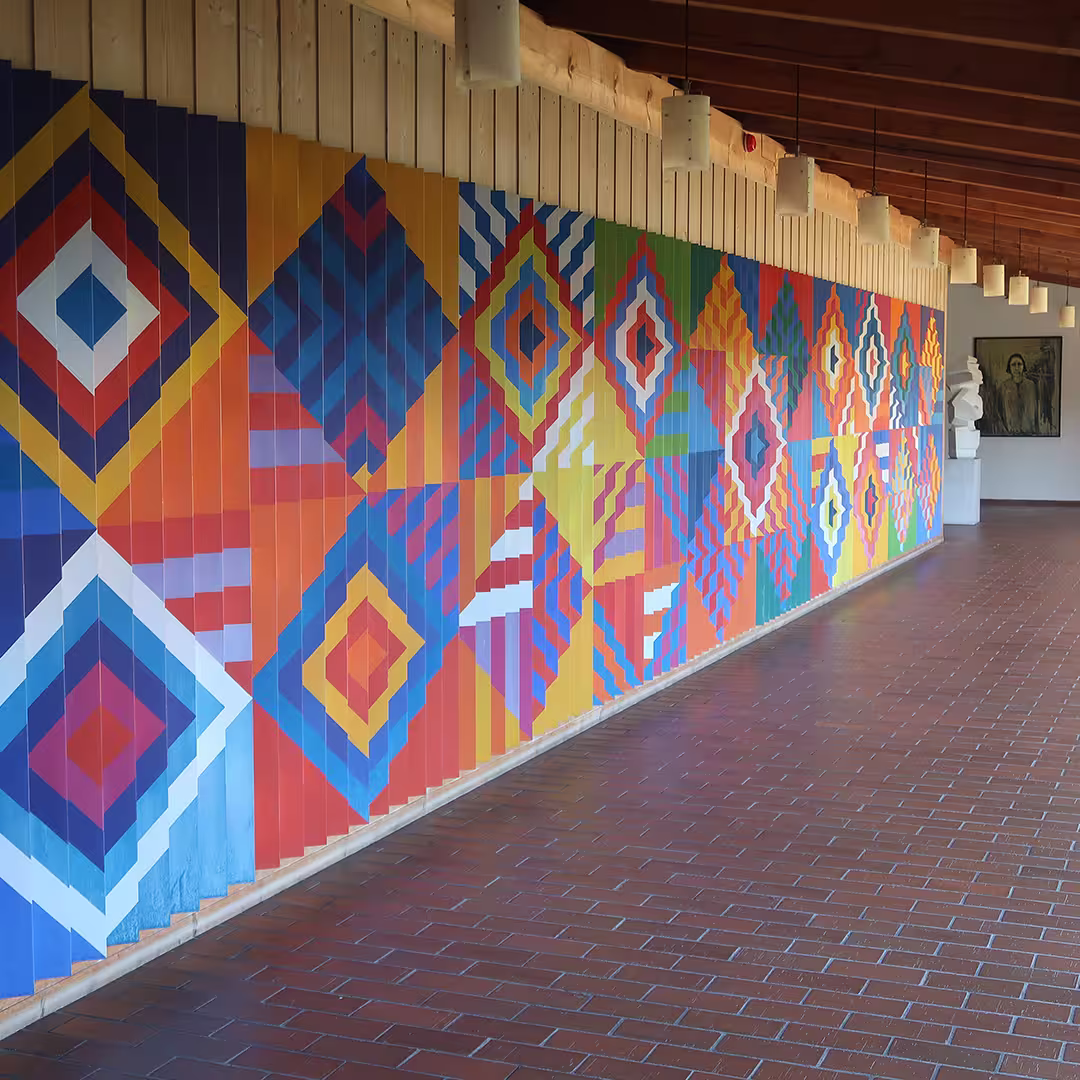 A long corridor at the Konventum Convention Centre in Elsinore, Denmark, featuring a striking geometric mural. The artwork consists of vibrant diamond-shaped patterns in red, blue, yellow, orange, and white, creating a visually dynamic effect along the wooden-paneled wall. The corridor has a brick floor and a wooden ceiling with hanging cylindrical lights. In the background, sculptures and framed artwork are displayed, adding to the artistic atmosphere of the space.