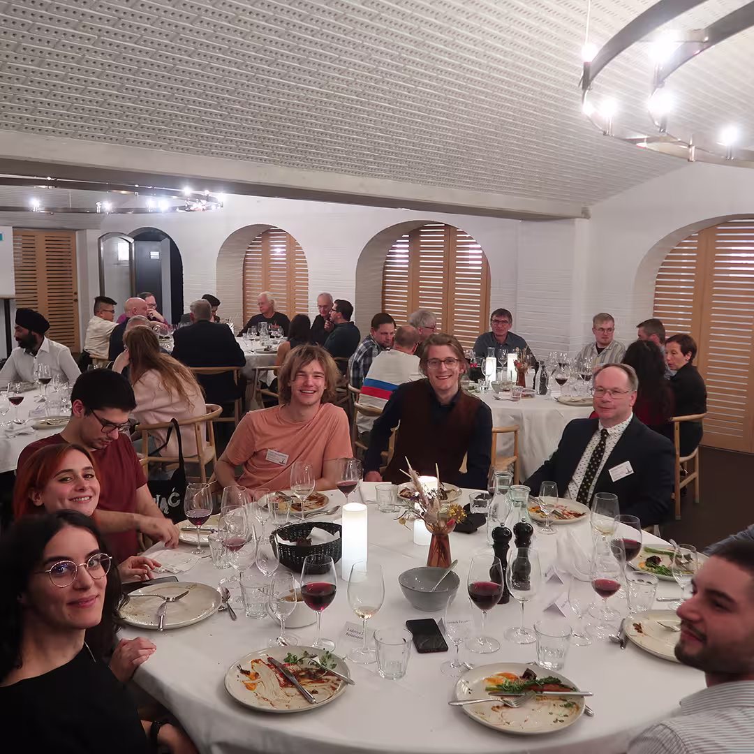 A lively dinner gathering at Dyalog '23, with attendees seated around a round table, enjoying conversation and laughter. The table is set with wine glasses, plates with partially eaten meals, and a decorative centerpiece with dried flowers. The group at the front includes individuals smiling towards the camera, with one person in a brown vest and another in a peach-colored shirt. Other tables in the background are filled with attendees engaged in discussions. The venue features arched windows with wooden shutters, a curved ceiling with perforated patterns, and modern lighting fixtures hanging overhead.
