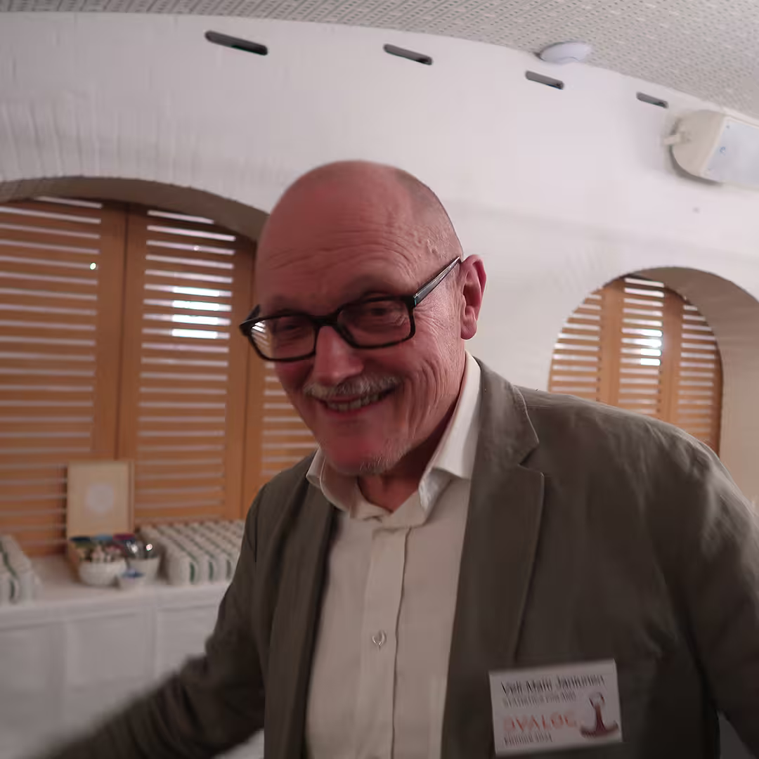 Veli-Matti Jantunen at Dyalog '23, smiling while wearing glasses, a white shirt, and a light olive-green blazer. His name badge is pinned to his jacket, displaying his name along with the Dyalog '23 logo. The background features a table with neatly arranged mugs and a bowl of condiments, set against arched windows with wooden shutters in a warmly lit room.