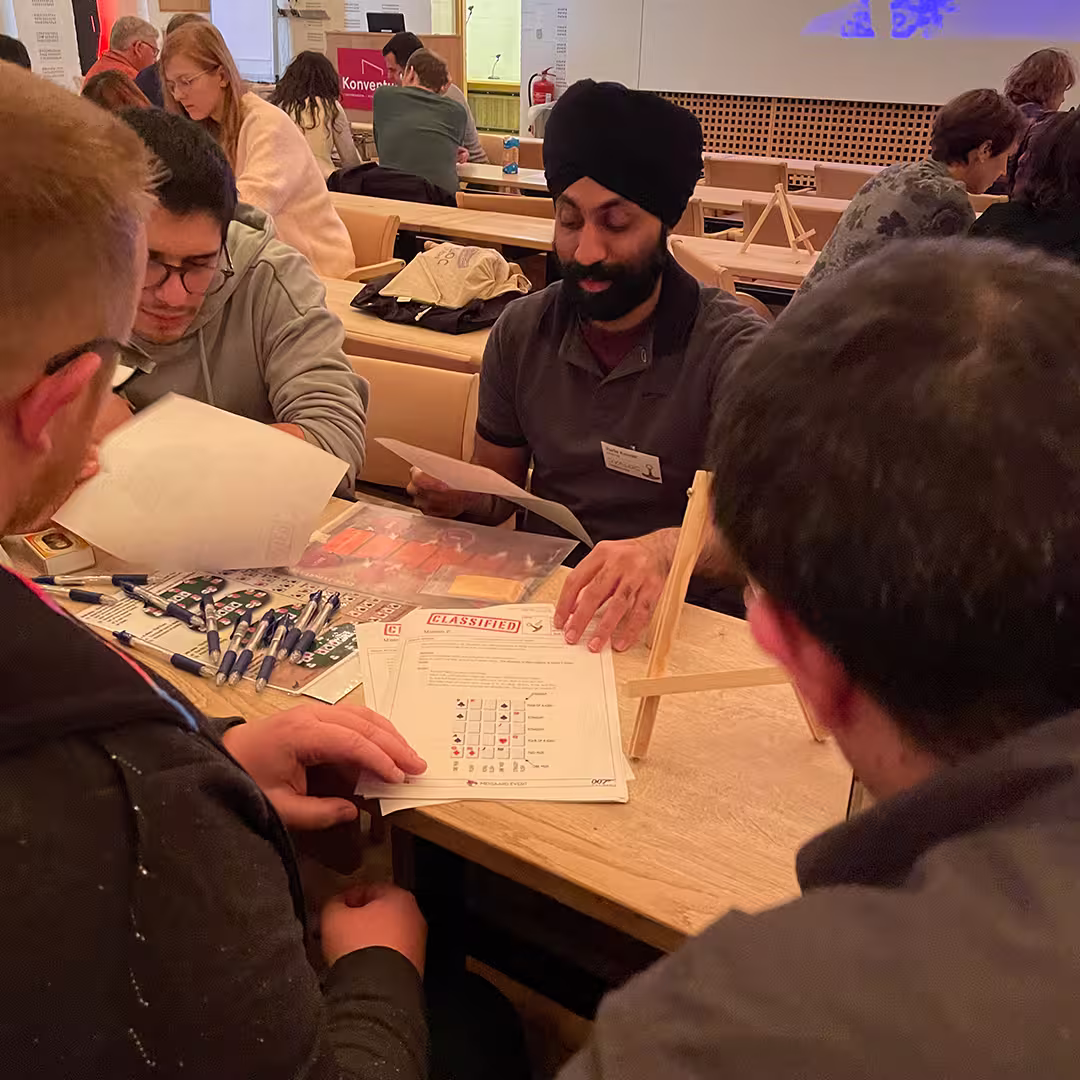 Karta Kooner, wearing a dark polo shirt and a black turban, participates in a group activity at Dyalog '23. He holds a sheet of paper while engaging in discussion with three other delegates seated around a wooden table. The table is covered with pens, documents labeled "CLASSIFIED," and game materials. In the background, other attendees are seated at tables in a warmly lit conference hall, with a presentation screen partially visible at the front of the room.