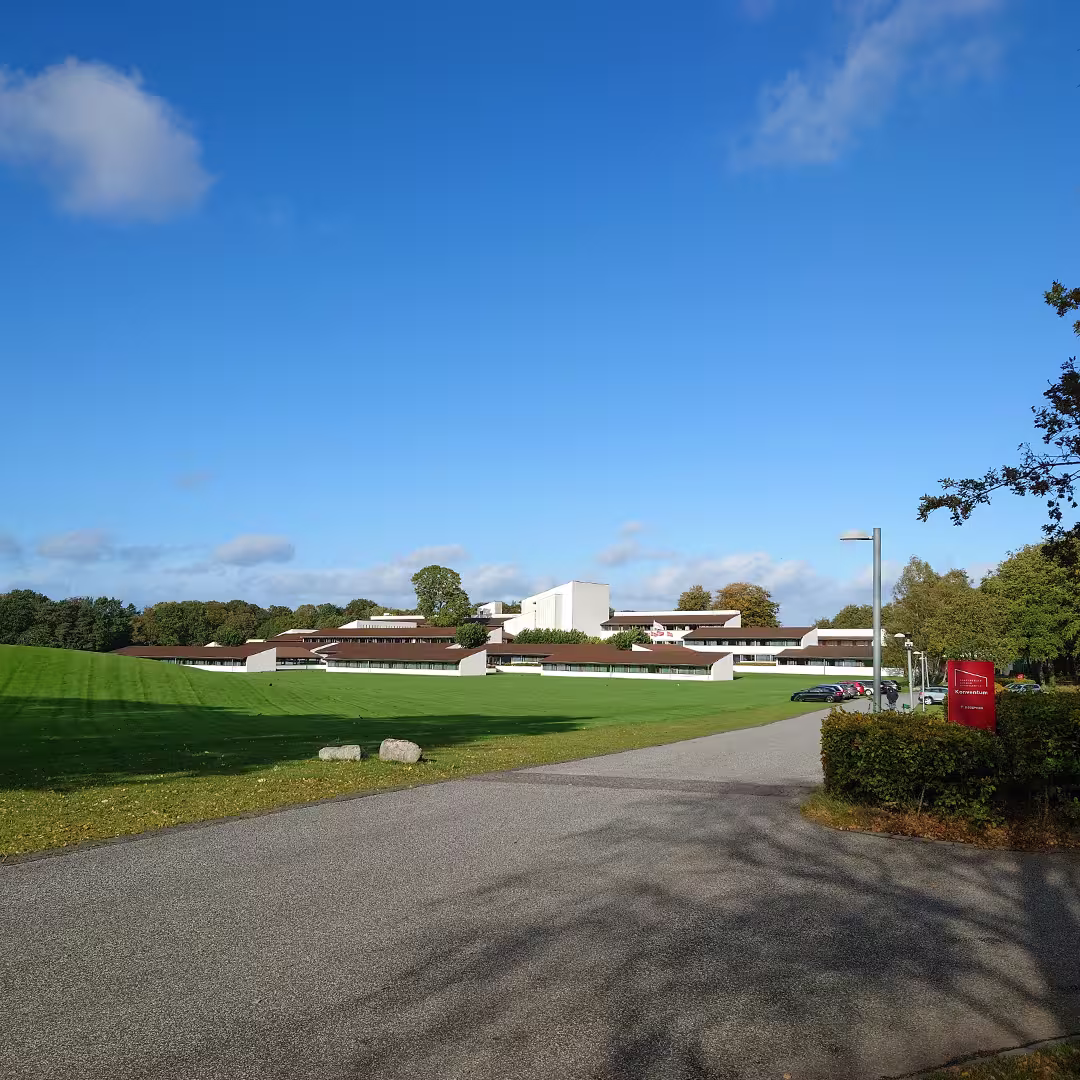 The Konventum Convention Centre in Elsinore, Denmark, the venue for Dyalog '23, surrounded by lush green lawns under a clear blue sky with scattered clouds. The modern, low-rise buildings have brown roofs and white walls, blending into the natural landscape. A paved driveway leads to the entrance, with a red signpost on the right. Several cars are parked in the distance, and trees line the background, adding to the serene setting.