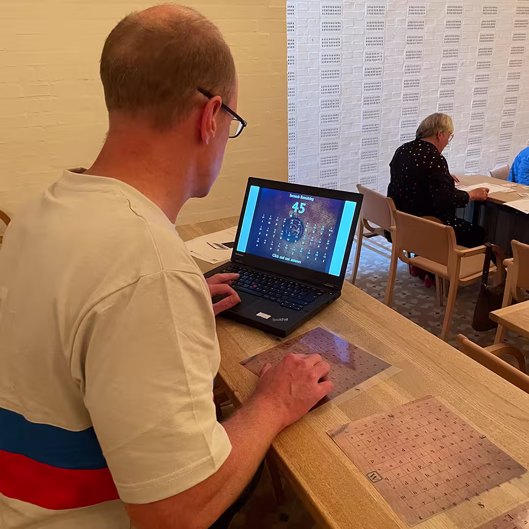 Stefan Kruger, wearing a white T-shirt with red and blue stripes, sits at a wooden table using a laptop at Dyalog '23. The screen displays a number puzzle game with a countdown timer. Transparent APL keyboard overlays are placed on the table in front of him. In the background, Gitte Christensen, wearing a black outfit with colorful dots, is seated at another table, engaged in an activity with papers in front of her.