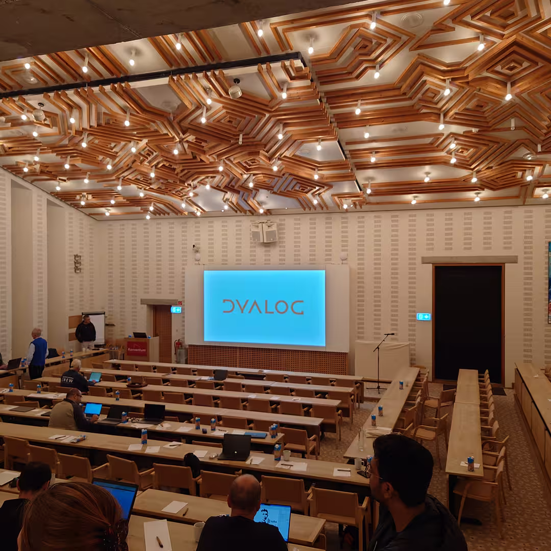 A lecture hall at Dyalog '23 with a large screen at the front displaying the Dyalog logo in blue. The room features tiered seating with long wooden tables, where several attendees are setting up laptops. The ceiling has an intricate geometric wooden pattern with numerous hanging light bulbs. A microphone stand is positioned near the stage, and a few people are gathered near the front, engaged in conversation. The walls are lined with a subtle textured pattern, contributing to the modern yet warm ambiance of the venue.