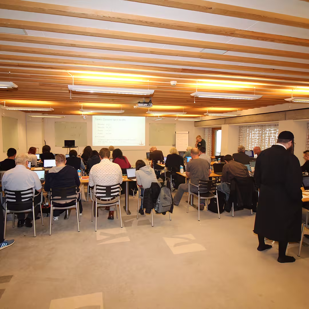 A workshop session at Dyalog '23 with attendees seated in rows, working on laptops. A presentation slide at the front displays a task titled "Convert text to dfns," with bullet points outlining the process. A presenter stands near a whiteboard, engaging with the audience. The room features wooden ceiling beams, soft lighting, and a modern layout. Some attendees are taking notes, while others listen attentively.
