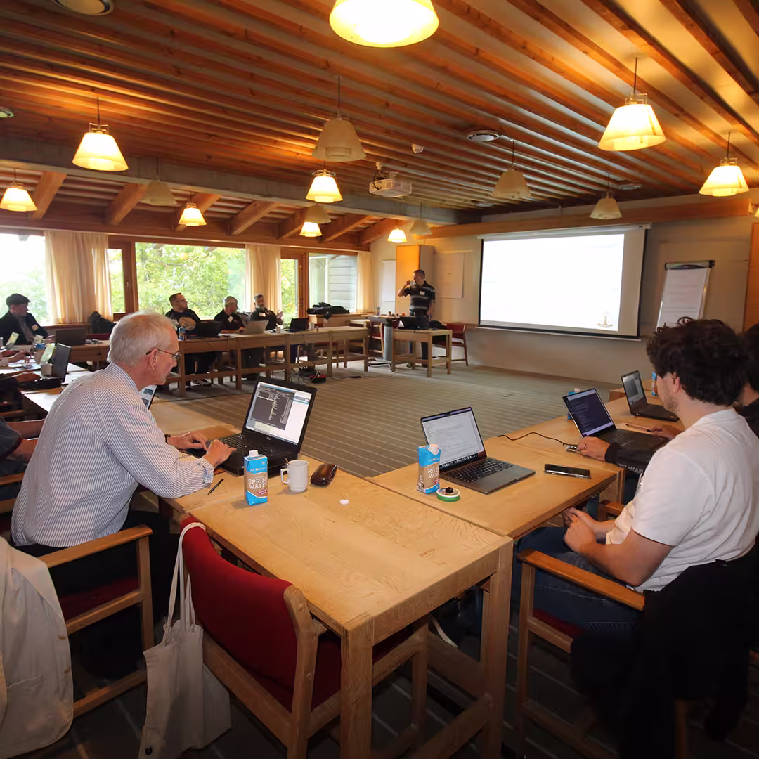 A workshop session at Dyalog '23 in a spacious conference room with a wooden ceiling and large windows allowing natural light to enter. Attendees, seated at wooden tables arranged in a U-shape, are working on laptops, some running APL code. A presenter stands at the front, delivering a talk while pointing at a projected slide on a large screen. Whiteboards with notes are visible in the background. The room has a warm ambiance with hanging lamps and an open view of greenery outside.