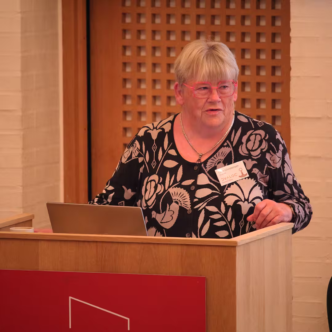 Gitte Christensen, wearing pink-framed glasses and a black cardigan with a bold floral and bird pattern, stands behind a wooden podium delivering a presentation at Dyalog '23. She has a name badge pinned to her cardigan and gestures with one hand while speaking. A laptop is open in front of her. The background features a wooden lattice panel and a white brick wall.