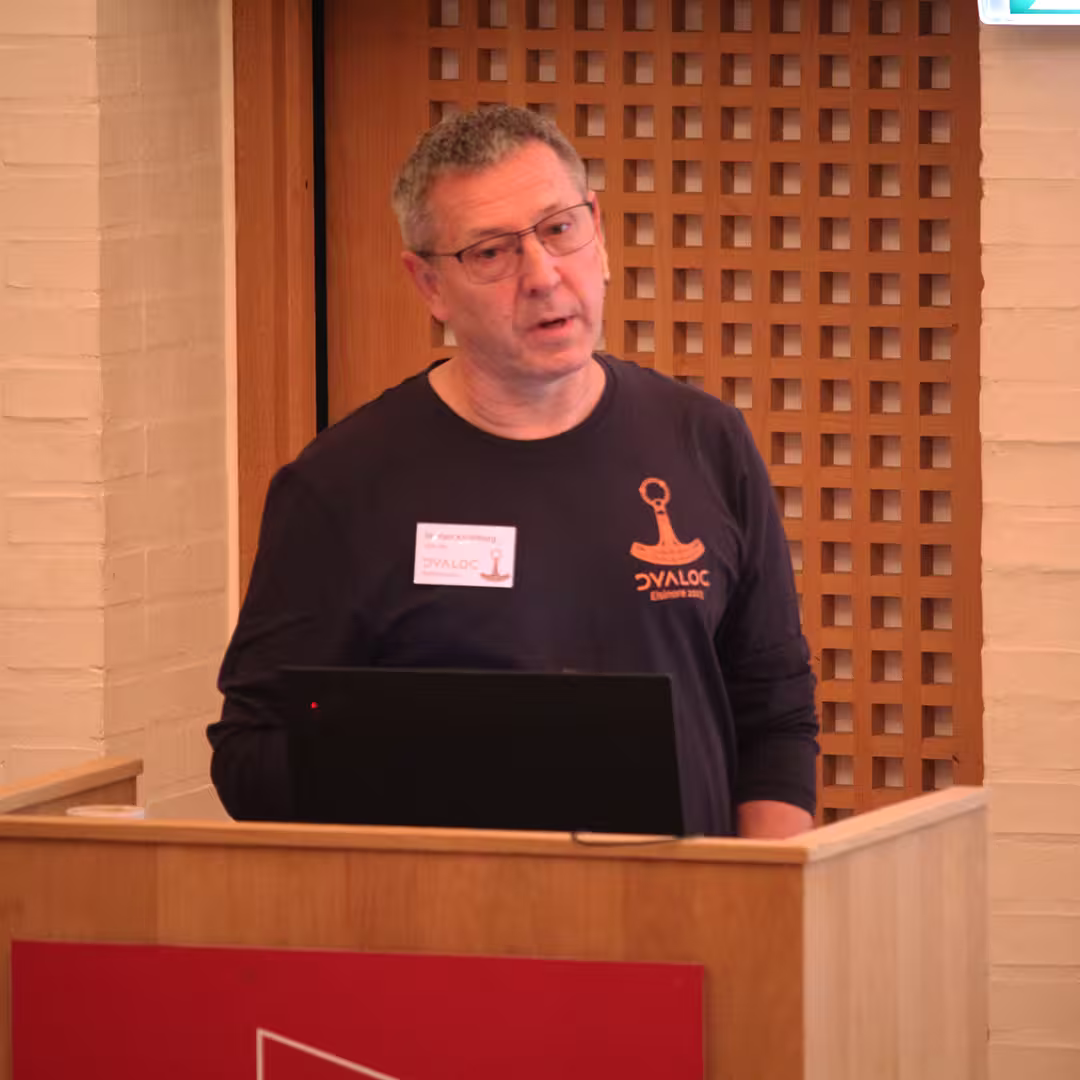 Morten Kromberg, wearing a black Dyalog '23 long-sleeve shirt with an orange APL logo, stands behind a wooden podium delivering a presentation. He has a name badge on his shirt and is speaking while looking towards the audience. A laptop is open in front of him. The background features a wooden lattice panel and a white brick wall.