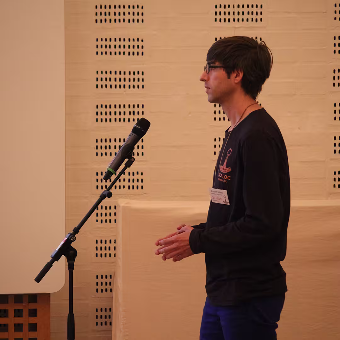 Brandon Wilson, wearing a black Dyalog '23 long-sleeve shirt with an orange APL logo, stands at a microphone asking a question during a session. His name badge is visible on his shirt. He gestures slightly with his hands as he speaks, looking towards the front of the room. The background features a white brick wall with ventilation holes and part of a projector screen.