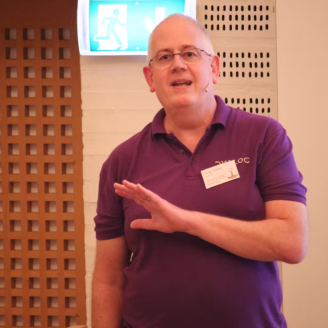 Andy Shiers, wearing a purple Dyalog polo shirt and a name badge, delivers a presentation at Dyalog '23. He is speaking while gesturing with his right hand. A headset microphone is positioned near his mouth. The background features a white brick wall, a wooden lattice panel, and an illuminated emergency exit sign above his head.