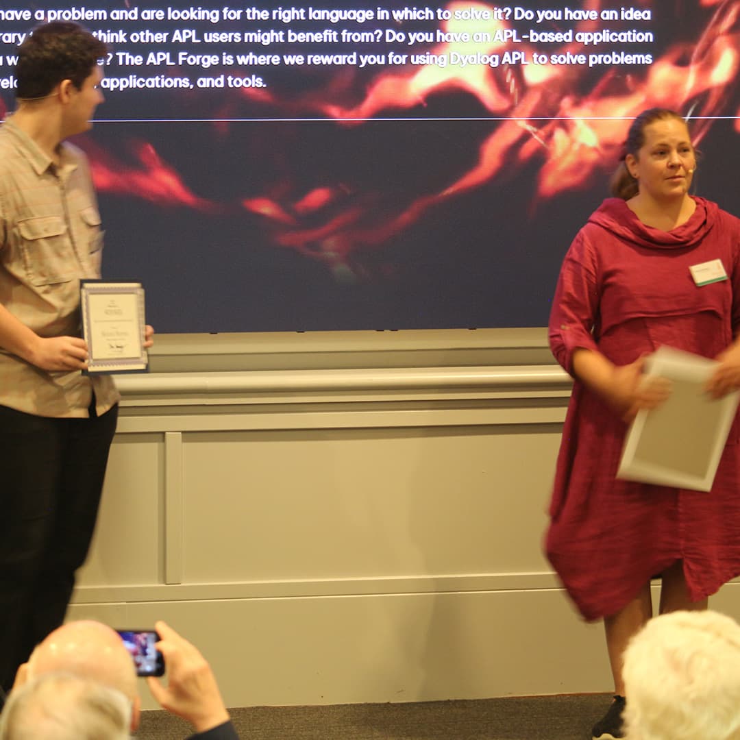 Holden Hoover and Stine Kromberg on stage during Dyalog '24 at the Golden Jubilee Hotel in Glasgow, Scotland. Holden, wearing a light-coloured plaid shirt, holds a framed award and looks towards Stine. Stine, dressed in a deep red dress, holds a certificate while speaking. Behind them, a large screen displays text about the APL Forge competition with a fiery background. In the foreground, an audience member captures the moment on a smartphone.