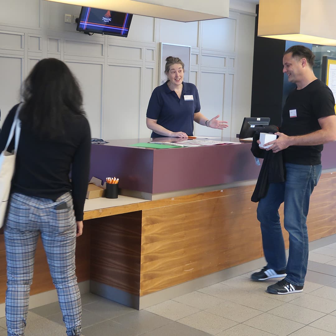Fiona Smith, wearing a navy blue polo shirt with a name badge, stands behind the registration desk at Dyalog '24, held at the Golden Jubilee Hotel in Glasgow, Scotland. She is gesturing while speaking to two delegates standing in front of the desk. One delegate, a woman with long dark hair, is seen from behind wearing a black top and plaid trousers, carrying a white tote bag. The other delegate, a man in a black t-shirt and jeans, holds a small white box and a jacket while smiling. The desk has pens, papers, and a monitor, with a screen above displaying the Dyalog logo.