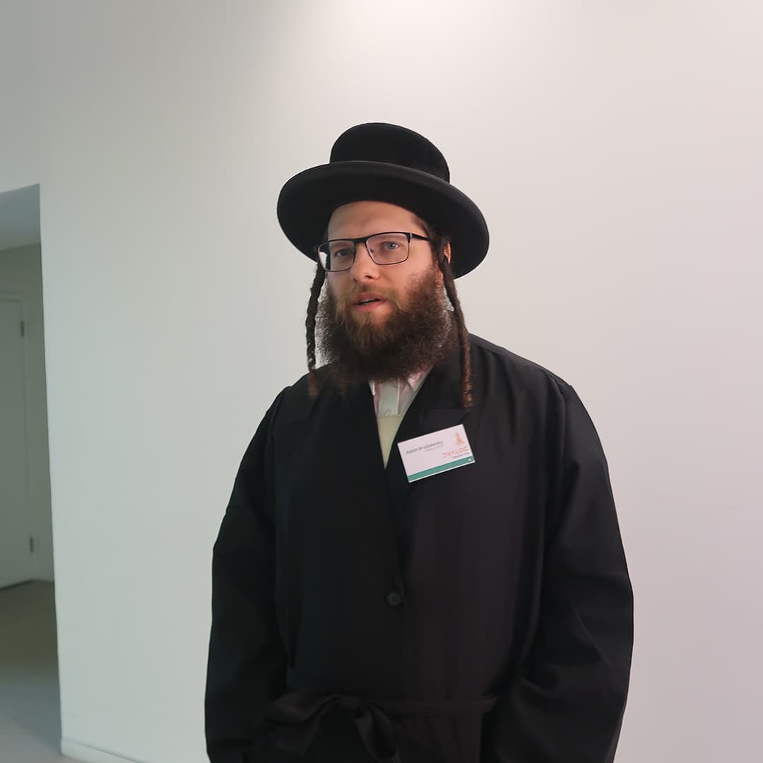 Adám Brudzewsky at Dyalog '24, held at the Golden Jubilee Hotel in Glasgow, Scotland. He is wearing a black coat and hat, along with glasses and a name badge. He stands against a plain white wall, looking towards the camera with a neutral expression. The setting appears to be a hallway or conference area with a doorway visible in the background.