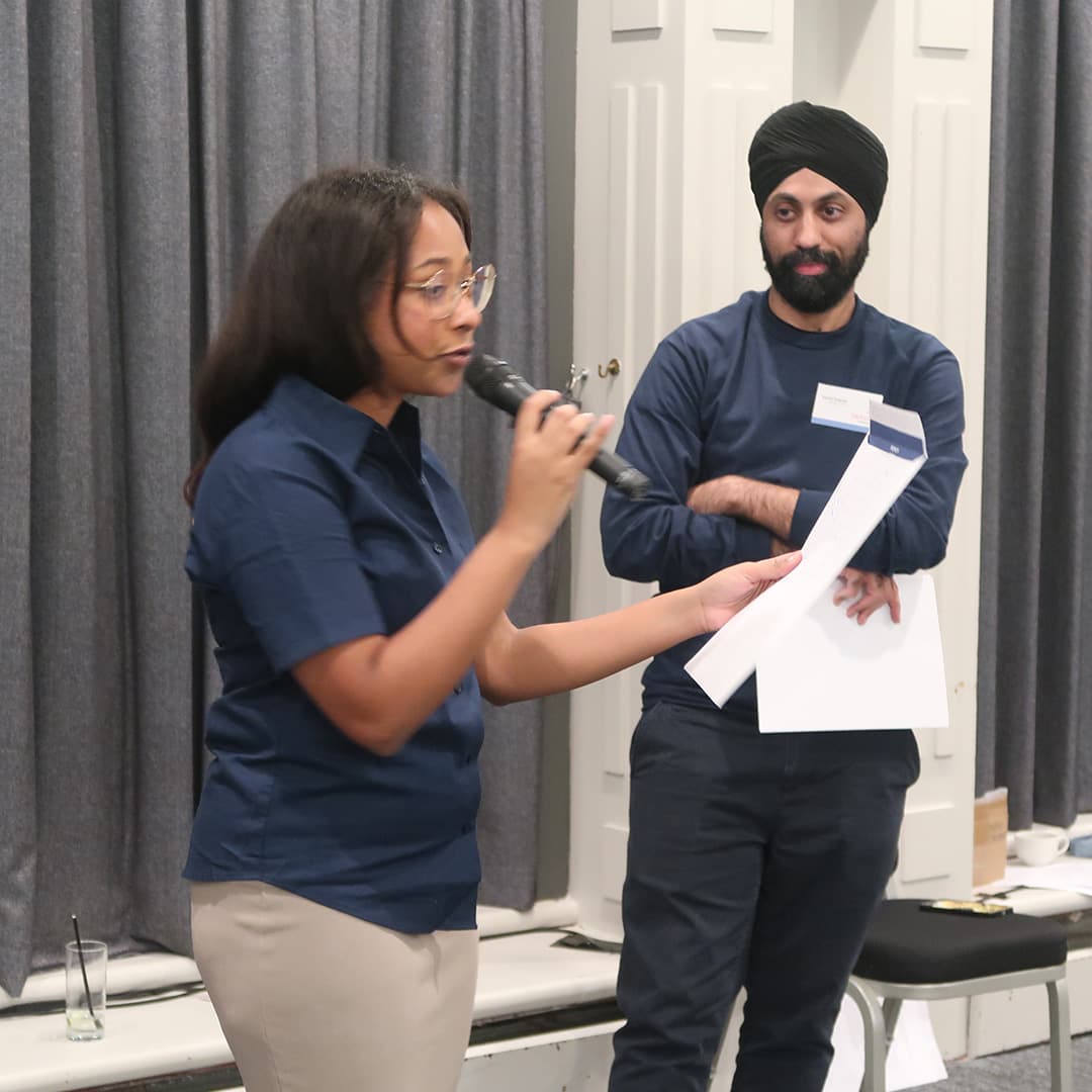 Jada Andrade and Karta Kooner hosting the Whyalog Quiz at Dyalog '24, held at the Golden Jubilee Hotel in Glasgow, Scotland. Jada, wearing a navy blue shirt and beige trousers, holds a microphone and a sheet of paper while speaking. Karta, also dressed in navy blue, stands beside her with arms crossed, holding additional papers. The background features grey curtains and white panelled walls, with a chair and various items scattered on the floor. A nearly empty glass with a straw sits on the stage edge.