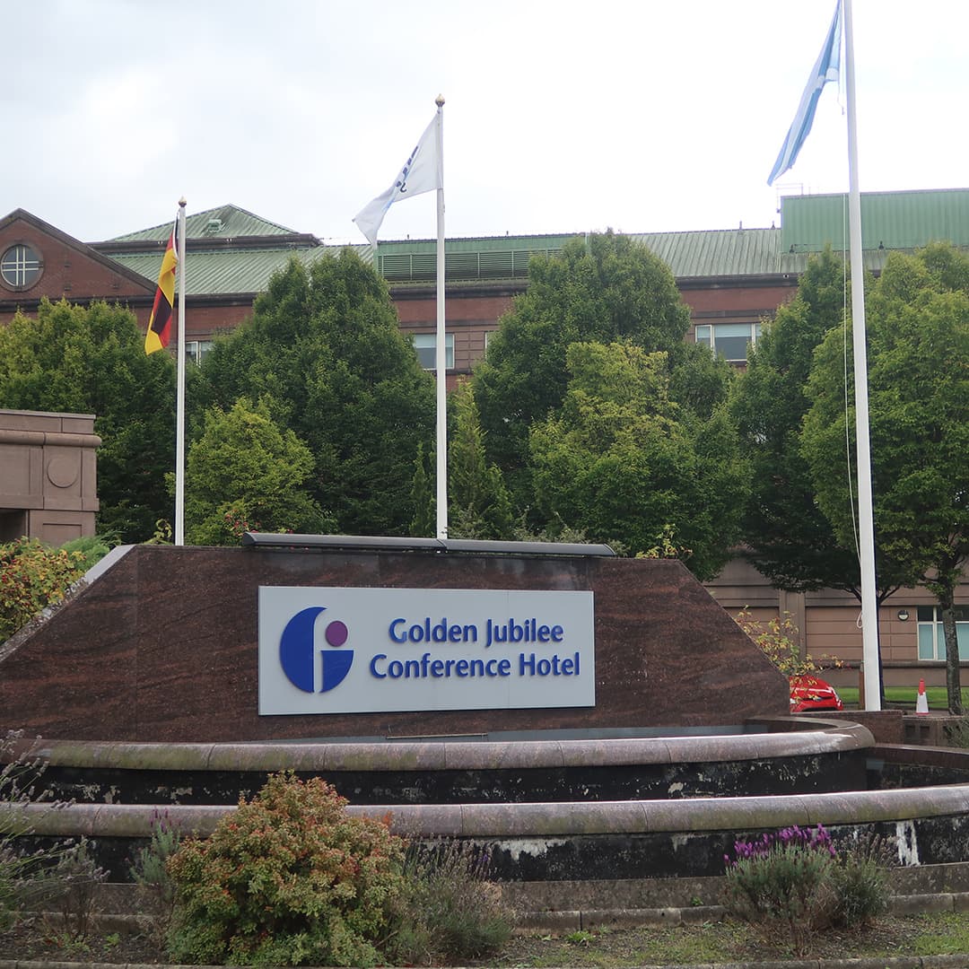 The entrance sign of the Golden Jubilee Conference Hotel in Glasgow, Scotland, the venue for Dyalog '24. The sign is mounted on a dark stone structure with a small water feature in front, surrounded by greenery and purple flowers. Three flagpoles stand behind the sign, flying the German, Dyalog, and Scottish flags. In the background, the hotel’s red brick and green-roofed building is partially visible, framed by lush trees.