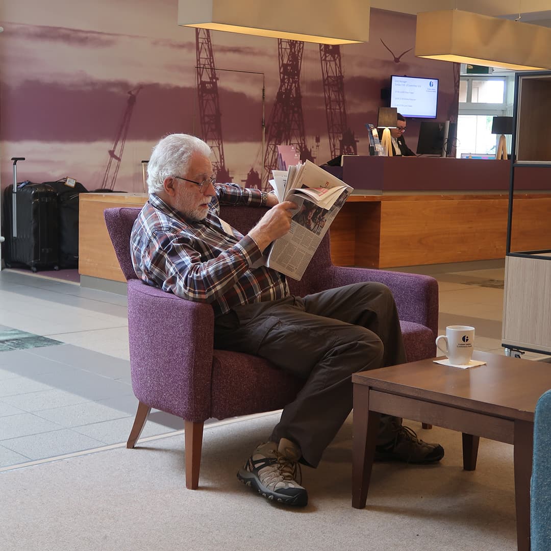 Geoff Streeter relaxing in the lobby of the Golden Jubilee Hotel during Dyalog '24 in Glasgow, Scotland. He sits in a purple armchair, wearing a plaid shirt and dark trousers, reading a newspaper. A cup of coffee with the hotel’s logo rests on a wooden table in front of him. In the background, the reception desk is visible with a staff member seated behind it, along with a large mural featuring oil rig silhouettes. A set of suitcases is positioned near the desk, suggesting recent arrivals or departures.