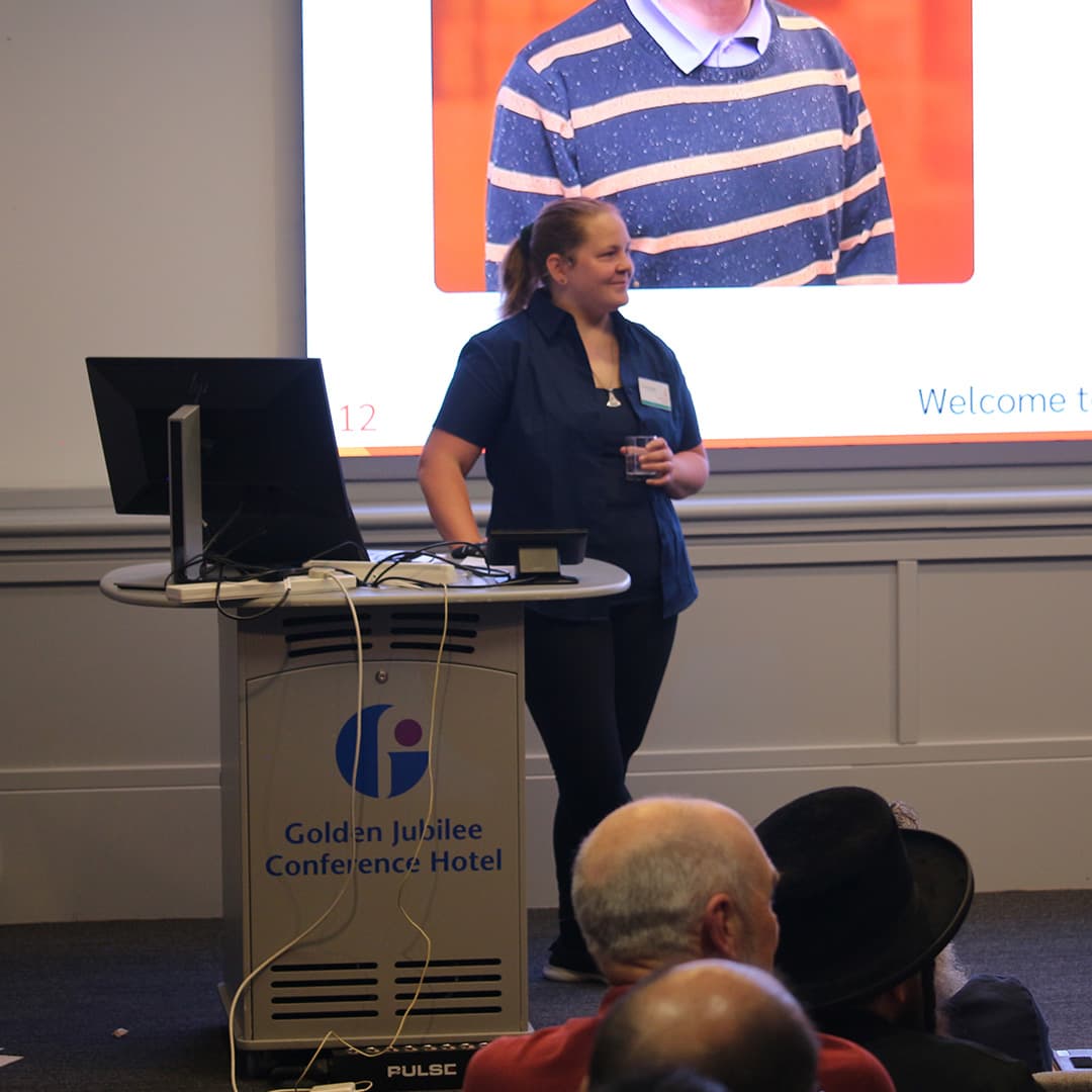 Stine Kromberg presenting at Dyalog '24, held at the Golden Jubilee Hotel in Glasgow, Scotland. She stands at the podium wearing a navy blue shirt, holding a glass of water and smiling. Behind her, a large screen displays a slide featuring a photo of another person and partially visible text. The podium is branded with the Golden Jubilee Conference Hotel logo, with cables and a monitor on top. In the foreground, attendees watch the presentation, including Adám Brudzewsky, who is wearing a black hat.