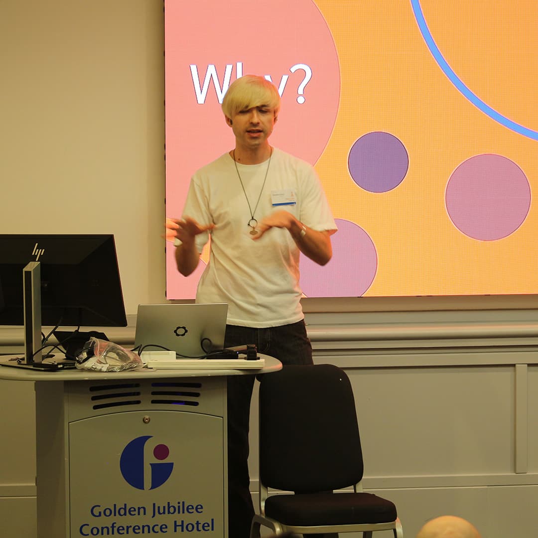 Brandon Wilson presenting at Dyalog '24, held at the Golden Jubilee Hotel in Glasgow, Scotland. He is wearing a white t-shirt and a pendant necklace, gesturing expressively while speaking. Behind him, a large screen displays a slide with an orange background, purple circles, and the word "Why?" in white text. In front of him, a podium with a laptop, a monitor, and some cables is branded with the Golden Jubilee Conference Hotel logo. A black chair is positioned beside the podium, and an attendee’s head is visible in the foreground.