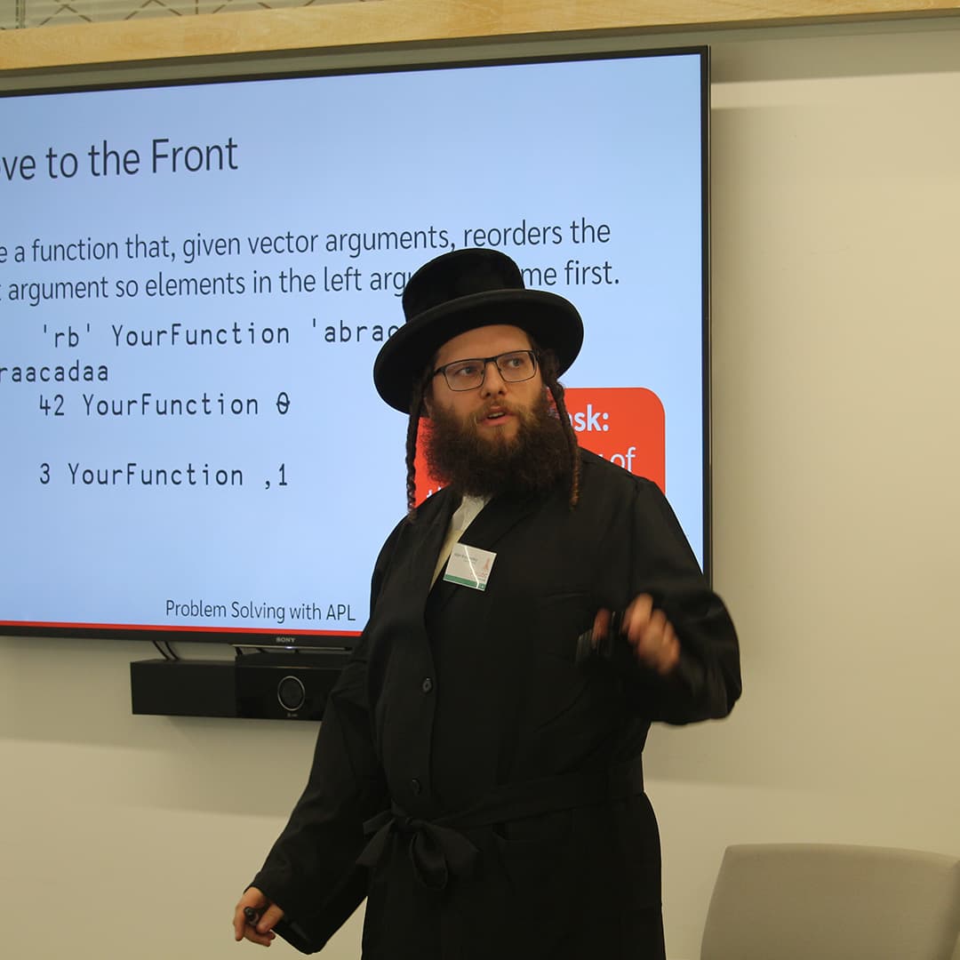 Adám Brudzewsky presenting at Dyalog '24, held at the Golden Jubilee Hotel in Glasgow, Scotland. He is wearing a black coat and hat, gesturing while speaking. Behind him, a large screen displays a slide titled "Move to the Front," featuring an APL coding challenge. The slide includes example code snippets and an instruction box with the text "Problem Solving with APL." A soundbar is mounted below the screen, and a grey chair is visible in the foreground.
