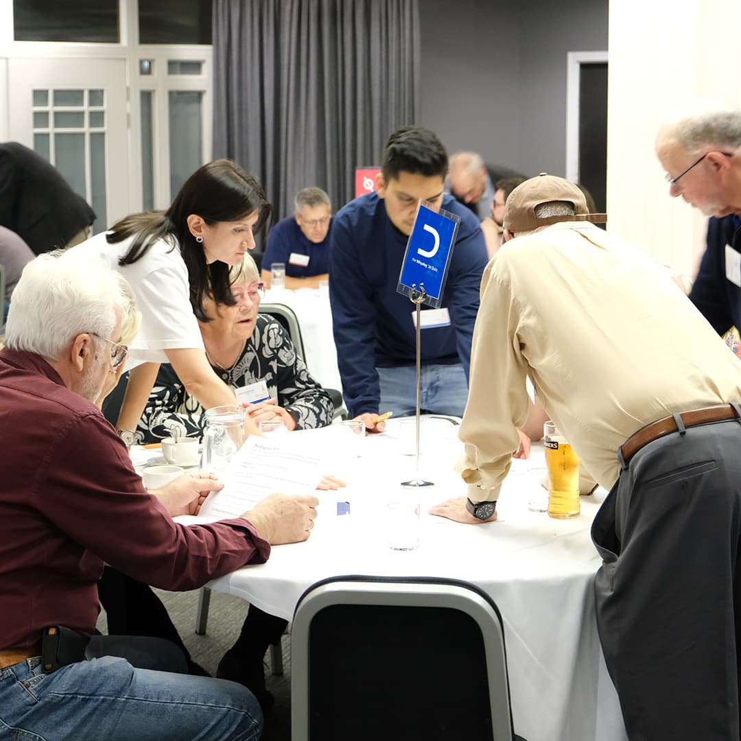 Attendees at Dyalog '24, held at the Golden Jubilee Hotel in Glasgow, Scotland, participate in the Whyalog Quiz. A group is gathered around a table marked with a blue sign featuring the Left Shoe (⊂) APL symbol. A woman in a white shirt leans in to discuss the answers with a seated attendee, while others stand or sit, engaged in conversation. Papers, glasses, and a pint of beer are scattered across the table. In the background, other quiz participants can be seen at their tables, with a red team sign visible further back. The room is well-lit, with dark curtains and large windows.