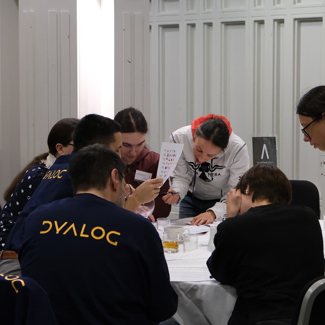 Attendees at Dyalog '24, held at the Golden Jubilee Hotel in Glasgow, Scotland, participate in the Whyalog Quiz. A group gathers around a table marked with a sign featuring the Lambda (∧) APL symbol. One attendee in a white hoodie leans in, pointing at a piece of paper filled with symbols, while others focus intently on the quiz. Several attendees wear navy blue Dyalog-branded shirts. The table is cluttered with papers, glasses, and a partially filled pint glass. The background features white panelled walls, and the room is brightly lit.