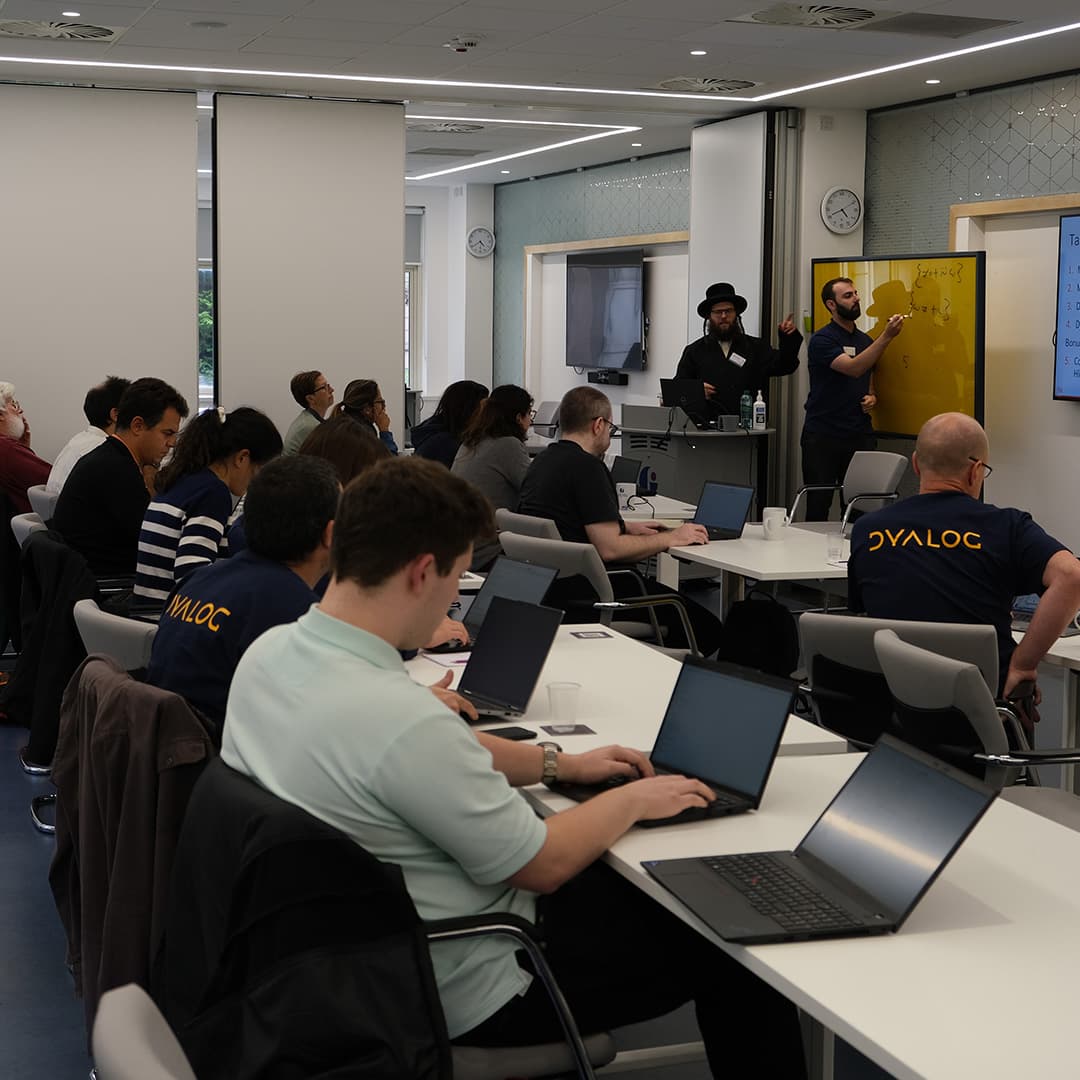 Adám Brudzewsky and Rich Park running a workshop at Dyalog '24, held at the Golden Jubilee Hotel in Glasgow, Scotland. Adám, wearing a black hat and dark clothing, stands near the front, gesturing while speaking. Rich, wearing a navy blue Dyalog polo shirt, writes on a yellow glass board. Attendees sit at tables with laptops open, taking notes or following along. Some wear Dyalog-branded shirts. The room is modern and well-lit, with large windows, white walls, and digital displays. A clock on the wall shows the time as the session progresses.