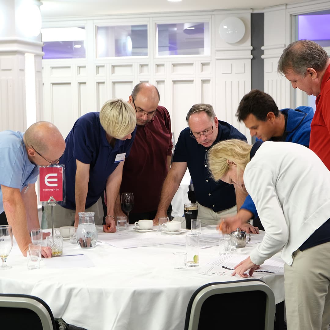 Attendees at Dyalog '24, held at the Golden Jubilee Hotel in Glasgow, Scotland, participate in the Whyalog Quiz. A group of seven attendees stands around a table, leaning in as they discuss their answers. The table is marked with a red sign featuring the Elements (∊) APL symbol. Papers, glasses, cups, and a bottle of Guinness are scattered across the table. The participants, dressed in a mix of casual and smart attire, appear focused as they work together on the quiz. The background features white panelled walls and soft lighting.