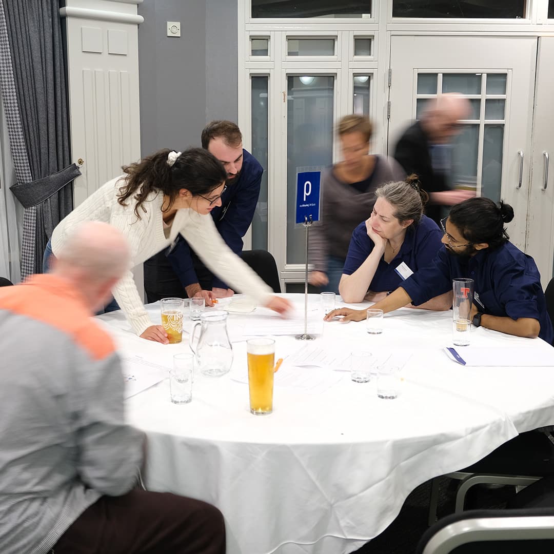 Attendees at Dyalog '24, held at the Golden Jubilee Hotel in Glasgow, Scotland, participate in the Whyalog Quiz. A group of attendees sits around a table marked with a blue sign featuring the Rho (⍴) APL symbol. A woman in a cream jumper leans forward, reaching across the table, while another woman (Fiona Smith) in a navy blue Dyalog shirt rests her chin on her hand, observing. A man with long dark hair and glasses gestures towards some papers. Drinks, papers, and glasses are scattered across the table. In the background, blurred figures move around the room, indicating an active and engaged atmosphere.