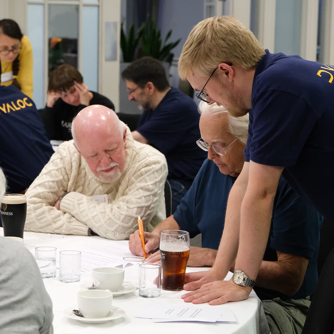 Attendees at Dyalog '24, held at the Golden Jubilee Hotel in Glasgow, Scotland, participate in a quiz. A young man wearing a navy blue Dyalog polo shirt leans over the table, assisting two older delegates. One delegate, wearing a cream-coloured cable knit jumper, listens intently, while the other, in a dark blue shirt and glasses, writes with an orange pen. The table is scattered with papers, glasses of water, a pint of beer, and cups of tea. In the background, other attendees are also engaged in the activity, with some wearing Dyalog-branded clothing.