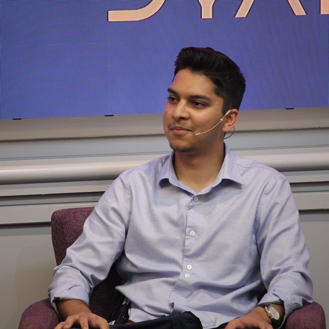 Josh David seated on stage during Dyalog '24 at the Golden Jubilee Hotel in Glasgow, Scotland. He is wearing a light blue button-up shirt and a headset microphone, appearing engaged in conversation. The background features a large screen displaying part of the Dyalog logo. He is sitting in a purple armchair with a relaxed posture, his hands resting on his lap.