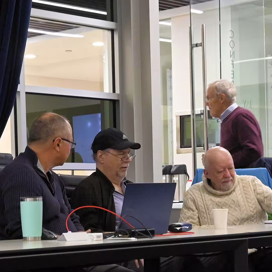 Three attendees seated at a table during DYNA Spring 2025 in New York, engaged with a laptop and conversation. The third person from the left, wearing a white jumper, is Robert Bernecky. Another person is visible walking past the glass doors in the background.