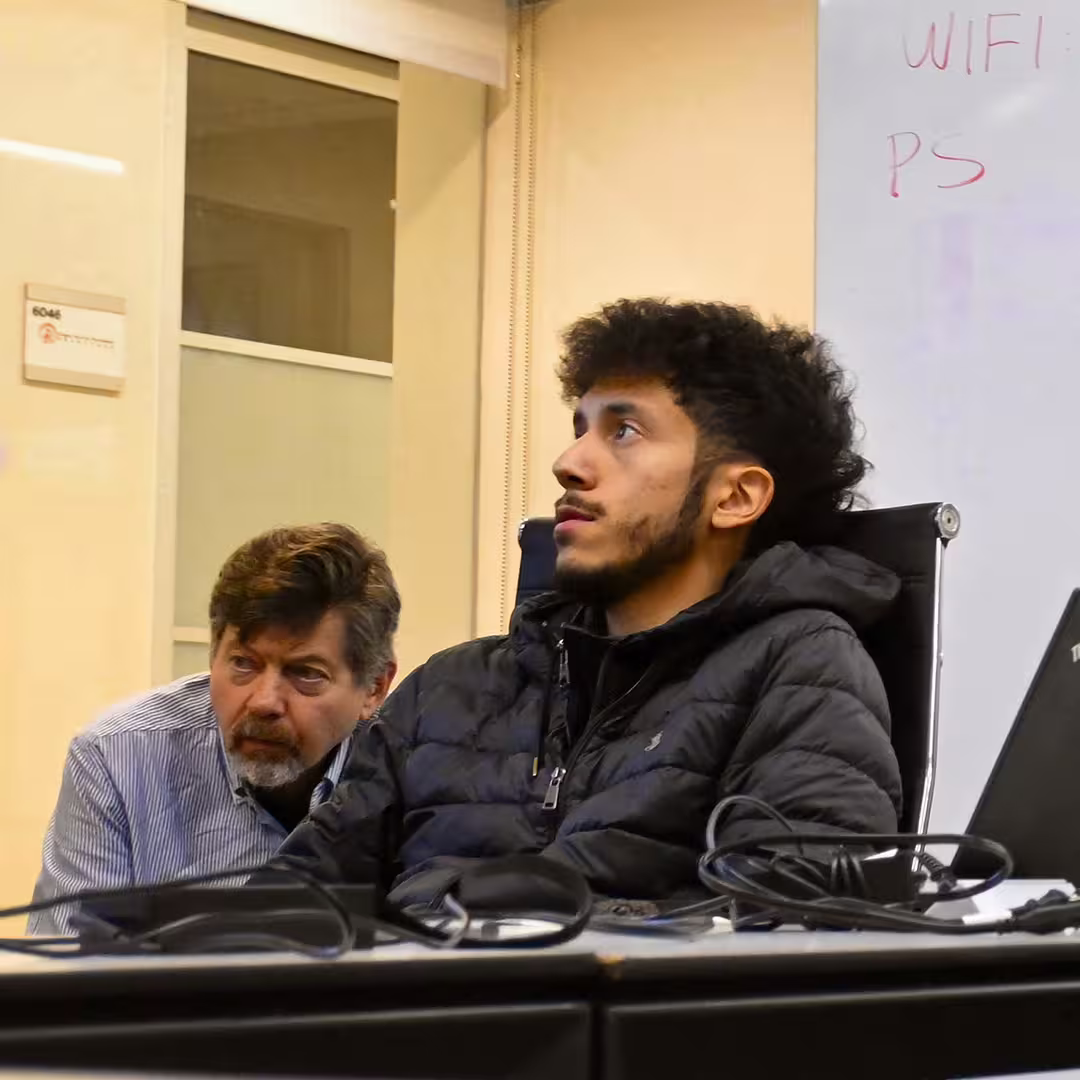 Mark Wolfson and Dexter Maldonado seated at a table during DYNA Spring 2025 in New York. Mark leans forward attentively while Dexter, wearing a black puffer jacket, looks ahead with focus. A laptop and cables are visible in the foreground.